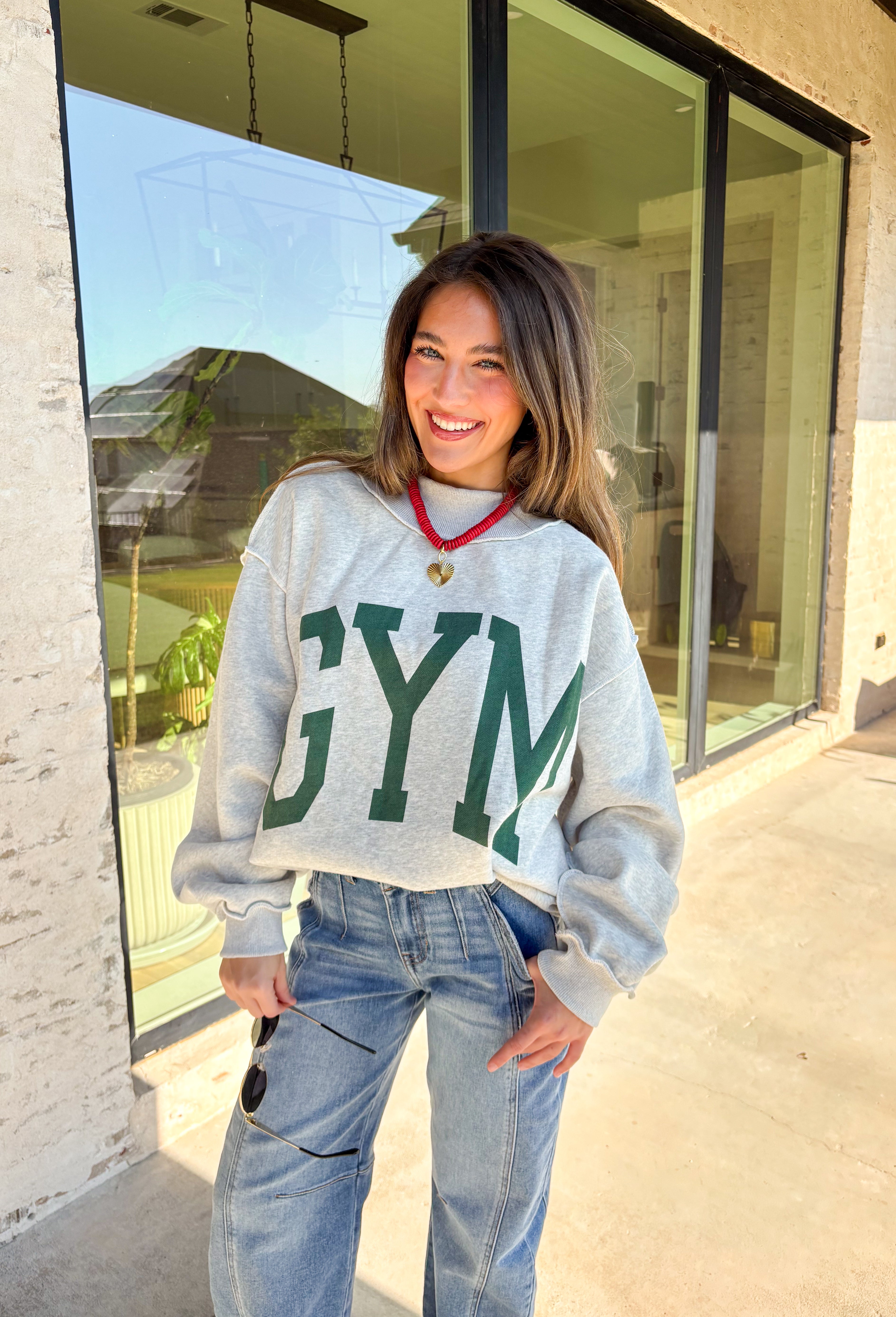 Two moods, one cozy pullover. This reversible mock neck sweater gives you options with light gray on one side and soft white fleece on the other. Printed in hunter green, one side reads “Gym” while the reverse says “Bed,” letting you wear your vibe for the day. With its oversized fit and cozy texture, it’s the perfect piece for lounging, layering, or heading out in casual comfort.