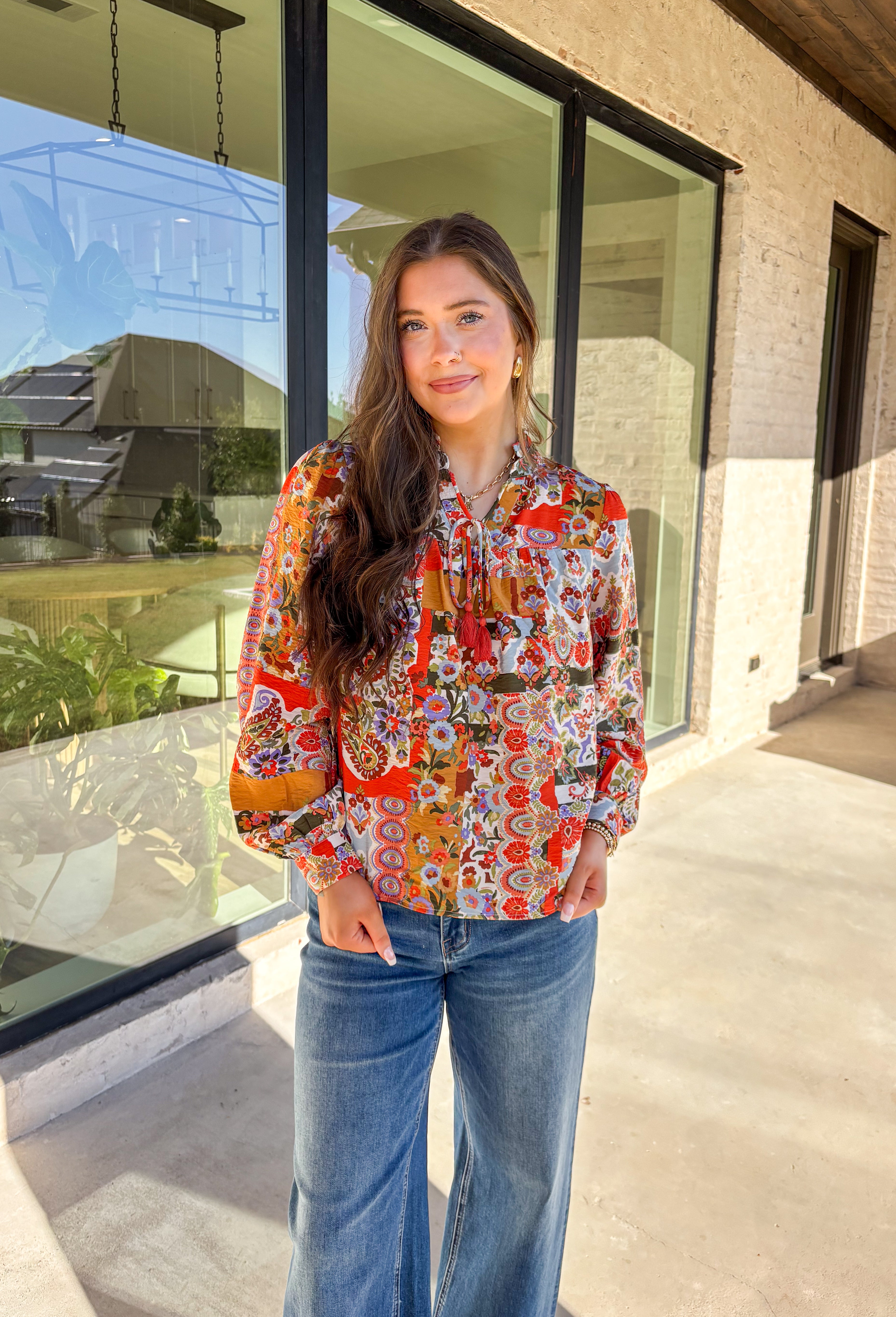 Bring bold color and cheerful energy to your fall wardrobe with this long sleeve floral top by Fate. The mix of orange, off white, tan, brown, purple, and blue creates a stunning statement print that feels both playful and timeless. Designed with a subtle ruffle neckline and a front tie detail, this blouse adds a soft feminine finish to your everyday look.