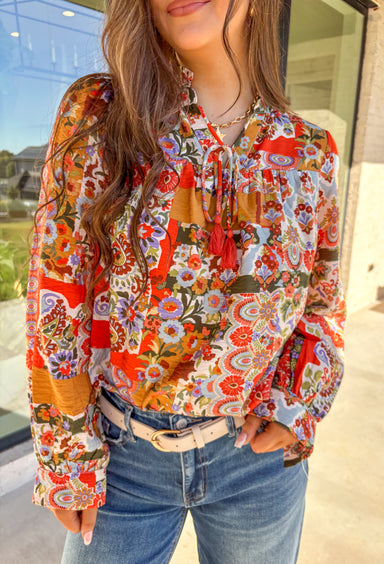 Bring bold color and cheerful energy to your fall wardrobe with this long sleeve floral top by Fate. The mix of orange, off white, tan, brown, purple, and blue creates a stunning statement print that feels both playful and timeless. Designed with a subtle ruffle neckline and a front tie detail, this blouse adds a soft feminine finish to your everyday look.