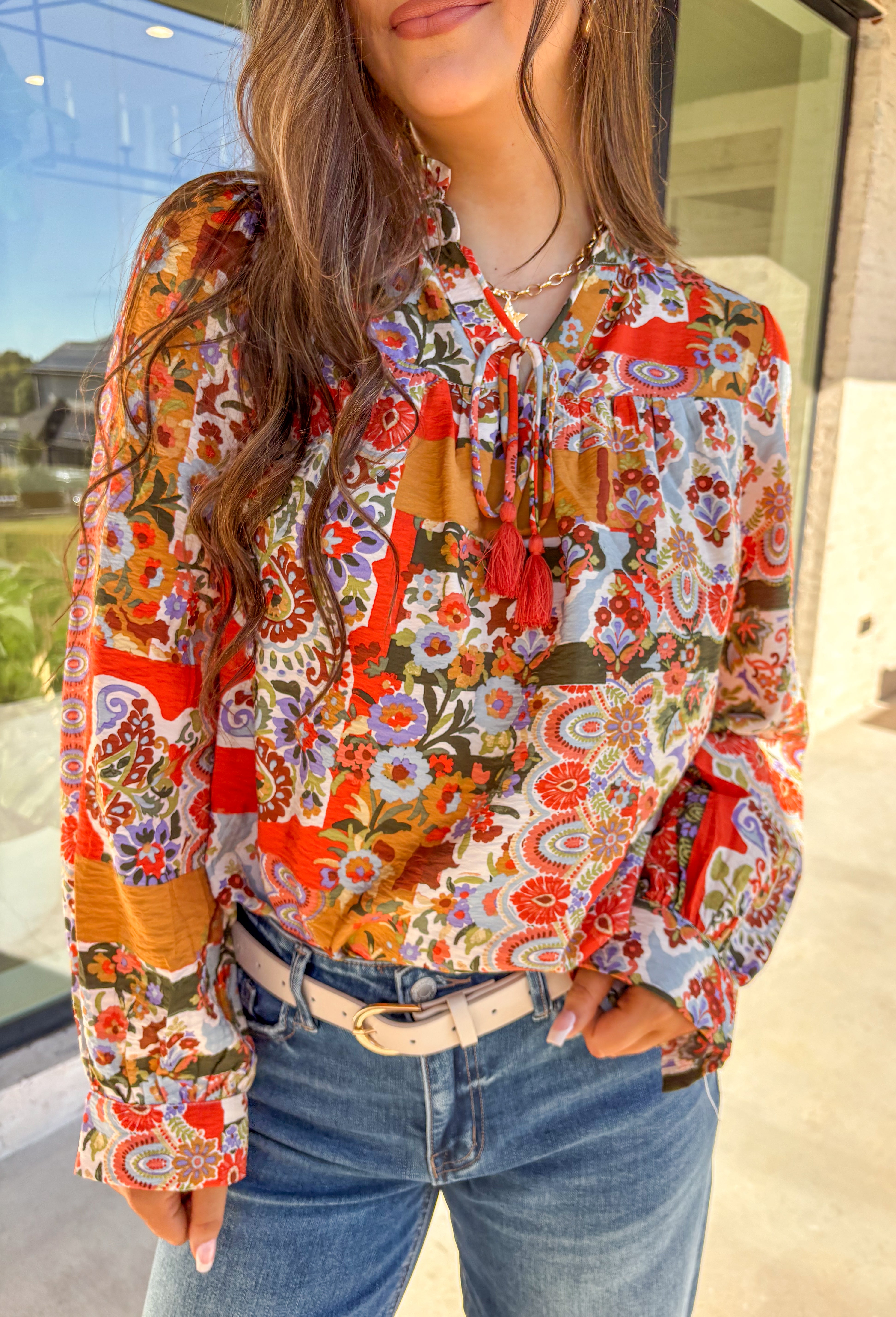 Bring bold color and cheerful energy to your fall wardrobe with this long sleeve floral top by Fate. The mix of orange, off white, tan, brown, purple, and blue creates a stunning statement print that feels both playful and timeless. Designed with a subtle ruffle neckline and a front tie detail, this blouse adds a soft feminine finish to your everyday look.