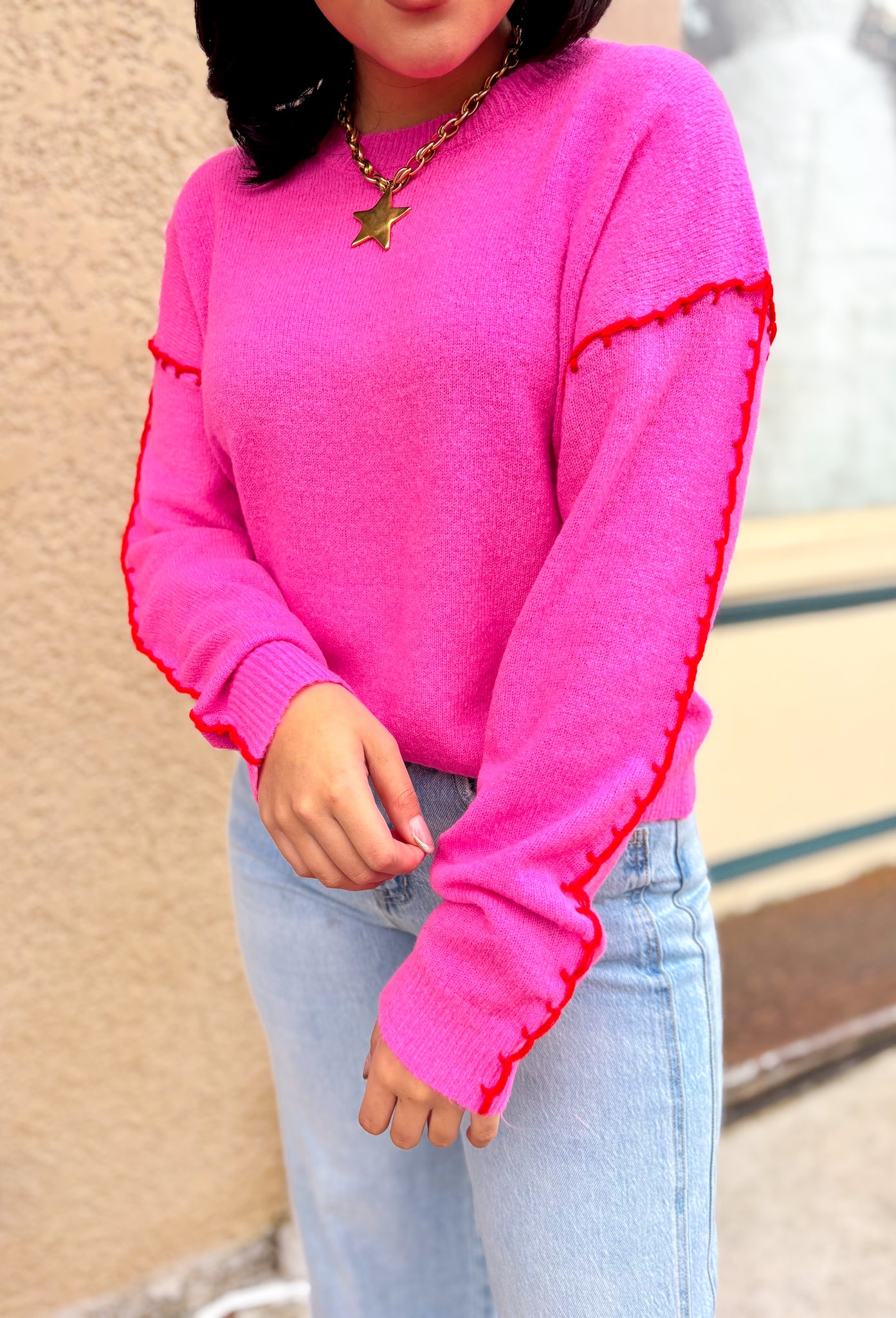 Pink sweater with a crew neck, long sleeves and a red whipstitch at shoulder stop and along top of sleeve. Styled with light wash jeans.