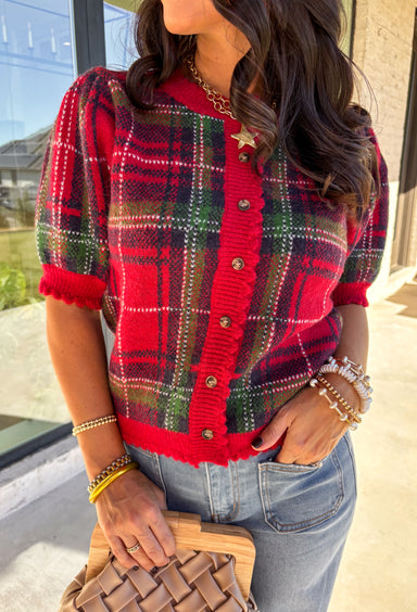 Festive and cozy, this plaid sweater knit top is made to bring holiday cheer. Featuring short puff sleeves and a scallop edge neckline, it’s finished with a button-front closure for a cardigan-style fit. The bold red plaid pattern in holiday tones makes it a standout for seasonal gatherings, while the soft knit keeps it comfortable and easy to wear. Perfect for layering or styling on its own, this cardigan blends playful charm with cozy comfort.
