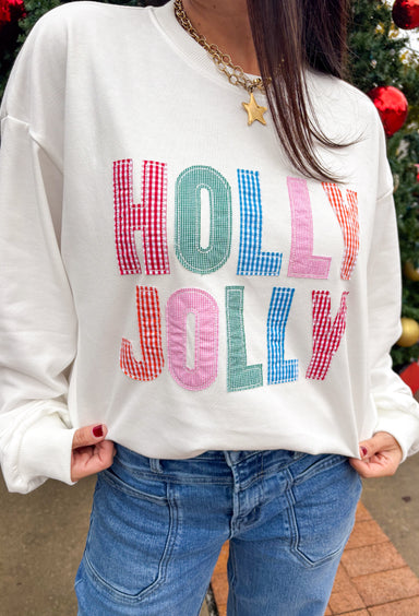 Stay merry and bright this season in the “Holly Jolly” pullover by Main Strip! This soft white lightweight sweatshirt features colorful gingham lettering—each letter a unique mix of festive color and pattern—outlined with white stitching for a playful holiday touch. Cozy and cheerful, it’s the perfect piece for decking the halls, sipping cocoa, or simply spreading a little Christmas joy.