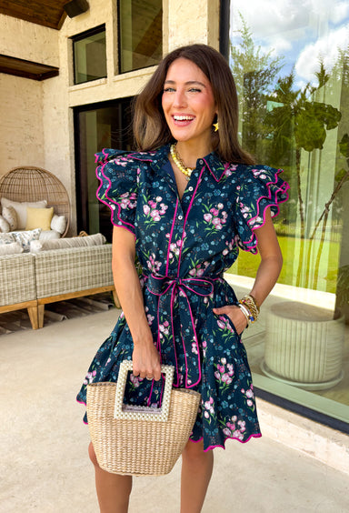 Bright, breezy, and made for spring plans, this navy dress by Entro is full of fun, feel-good color. Featuring a vibrant floral print with pops of purple, pink, green, and blue, it brings the perfect burst of spring energy to a classic navy base. The collared neckline and full button front give it a polished look, while flutter sleeves keep it light and feminine. Finished with contrast scallop trim along the edges, a waist tie for a flattering fit, and side pockets.