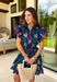 Bright, breezy, and made for spring plans, this navy dress by Entro is full of fun, feel-good color. Featuring a vibrant floral print with pops of purple, pink, green, and blue, it brings the perfect burst of spring energy to a classic navy base. The collared neckline and full button front give it a polished look, while flutter sleeves keep it light and feminine. Finished with contrast scallop trim along the edges, a waist tie for a flattering fit, and side pockets.