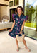Bright, breezy, and made for spring plans, this navy dress by Entro is full of fun, feel-good color. Featuring a vibrant floral print with pops of purple, pink, green, and blue, it brings the perfect burst of spring energy to a classic navy base. The collared neckline and full button front give it a polished look, while flutter sleeves keep it light and feminine. Finished with contrast scallop trim along the edges, a waist tie for a flattering fit, and side pockets.