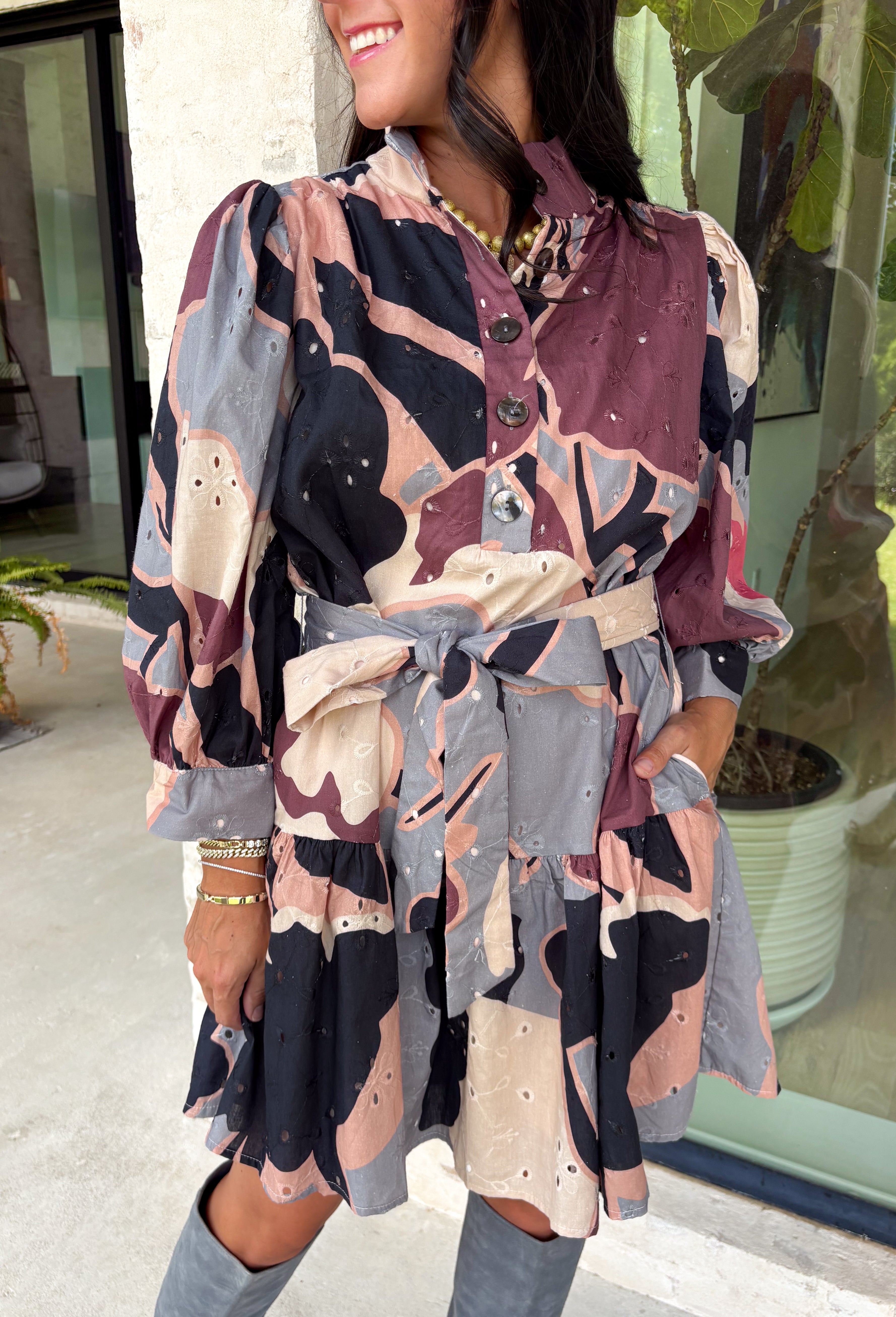 Celebrate the beauty of the season in this stunning eyelet dress from Entro. Designed in a gorgeous fall-inspired print featuring shades of black, brown, tan, gray, off-white, and a subtle pop of pink, this timeless dress is full of feminine charm. Delicate eyelet detailing adds beautiful texture, while the decorative front button accents and matching self-tie belt create a polished, flattering silhouette. 
