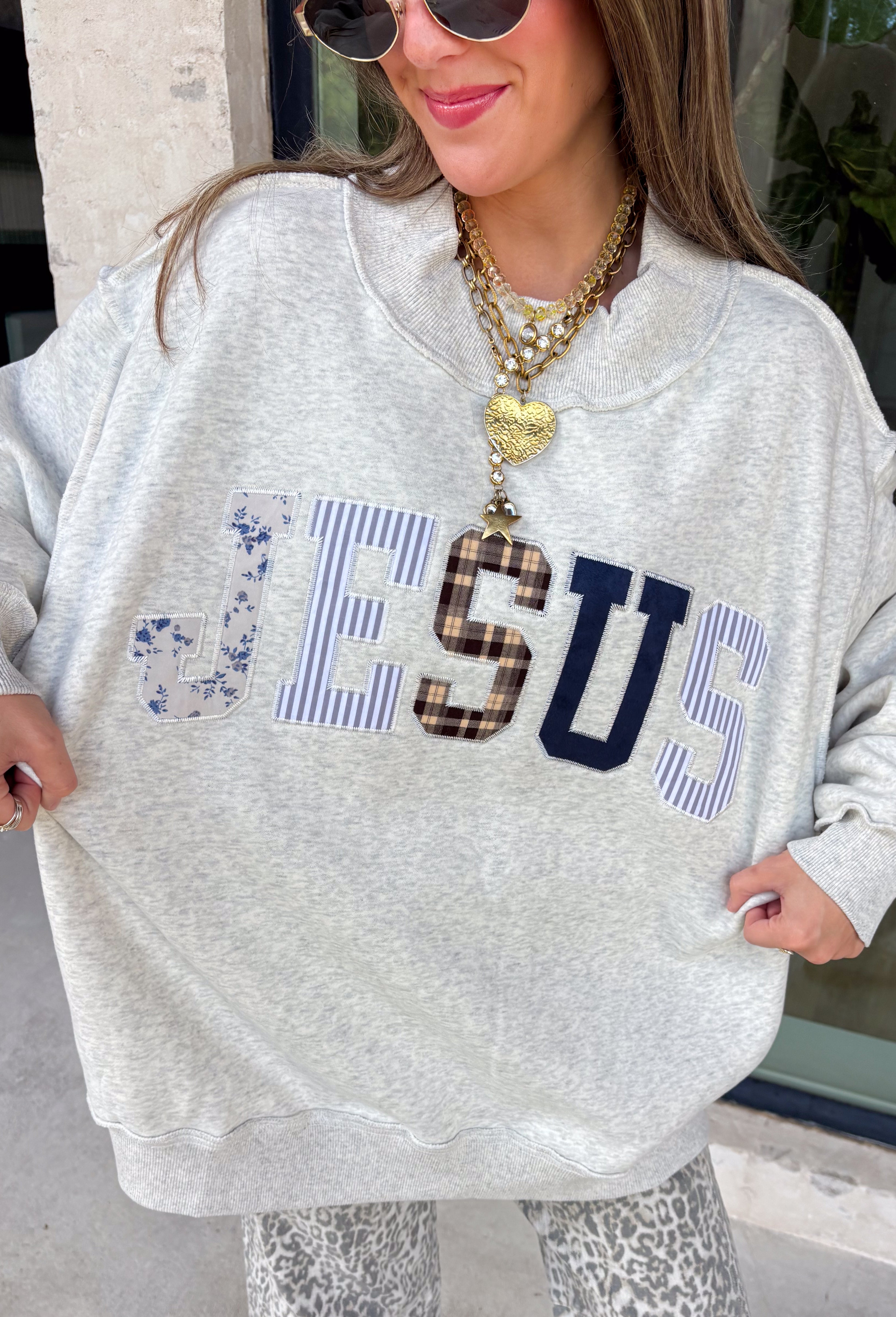 Wear your faith in the coziest way with this heather grey “Jesus” pullover from Bucket List. Each letter features its own unique print, including dainty florals, classic stripes, plaid, and solid navy in a soft mix of neutrals and shades of blue. Light grey embroidery outlines each letter for an extra touch of texture that makes the design stand out beautifully against the heather grey background. 