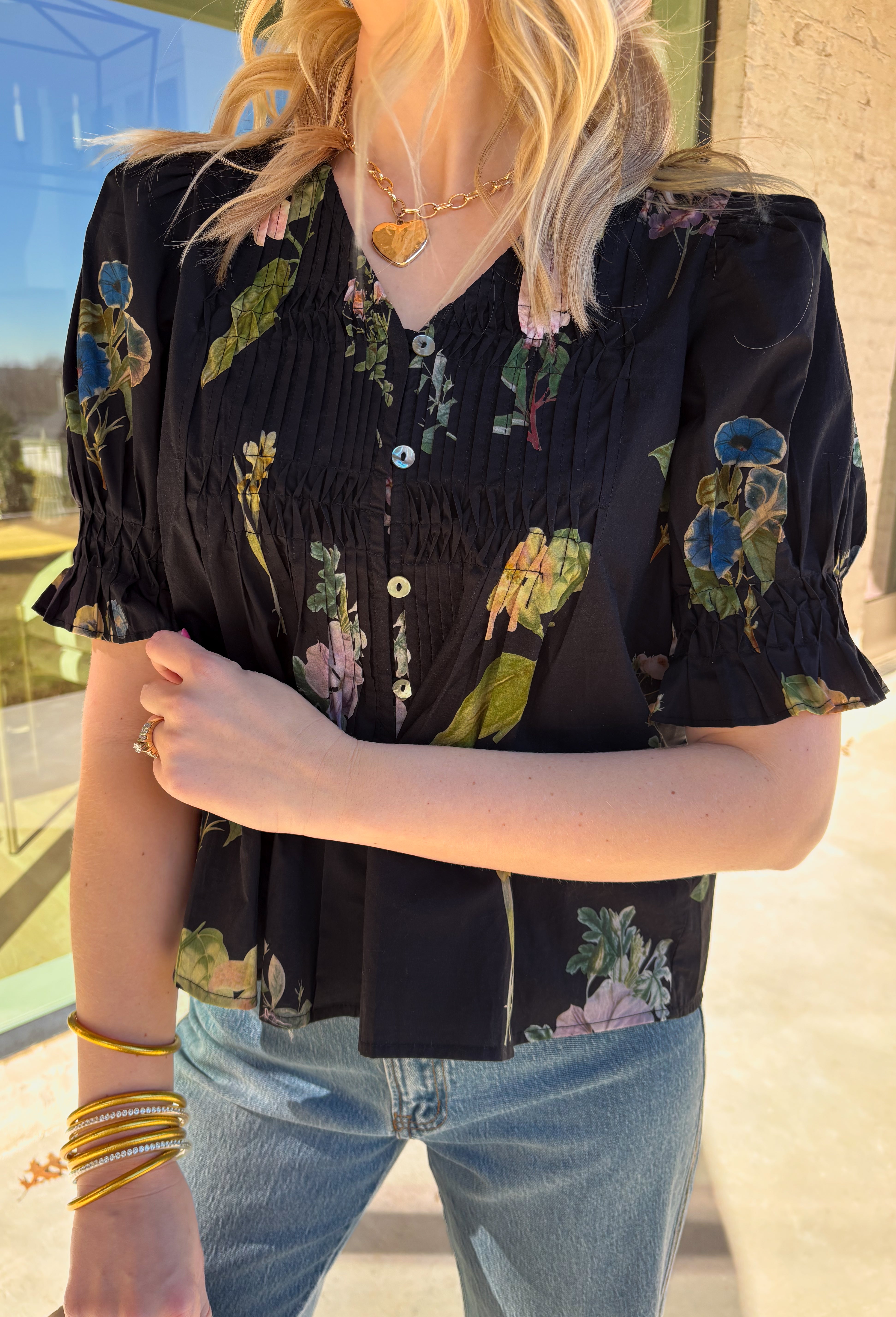 Classic with a spring refresh, this black short sleeve top by Entro is an easy standout. Featuring a soft floral print with pops of blush, green, and blue, it adds just the right amount of color to a timeless silhouette. The button-down front and pleating detail along the front give it a polished, flattering look, while the lightweight cotton fabric keeps it breathable and comfortable for warmer days.