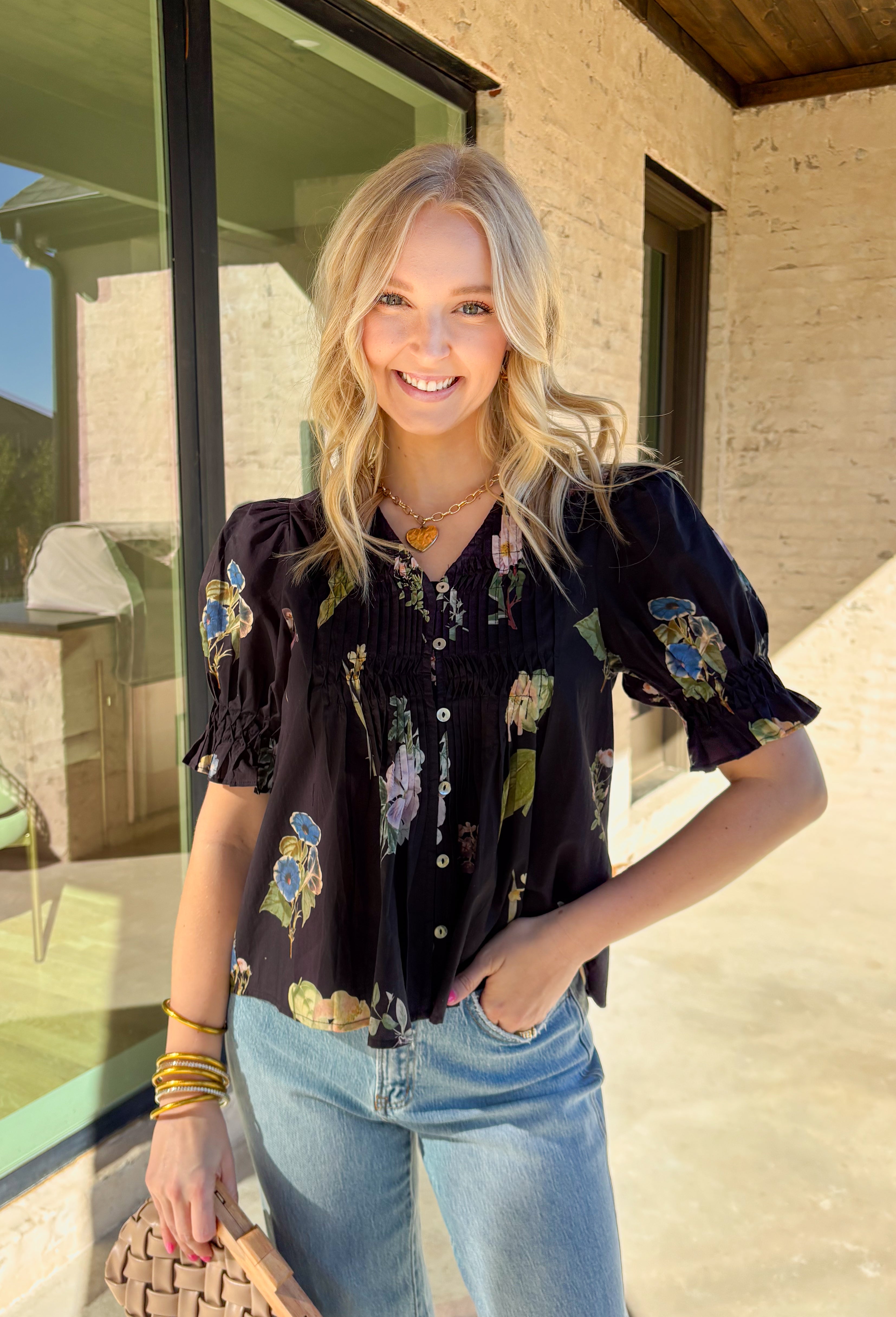 Classic with a spring refresh, this black short sleeve top by Entro is an easy standout. Featuring a soft floral print with pops of blush, green, and blue, it adds just the right amount of color to a timeless silhouette. The button-down front and pleating detail along the front give it a polished, flattering look, while the lightweight cotton fabric keeps it breathable and comfortable for warmer days.