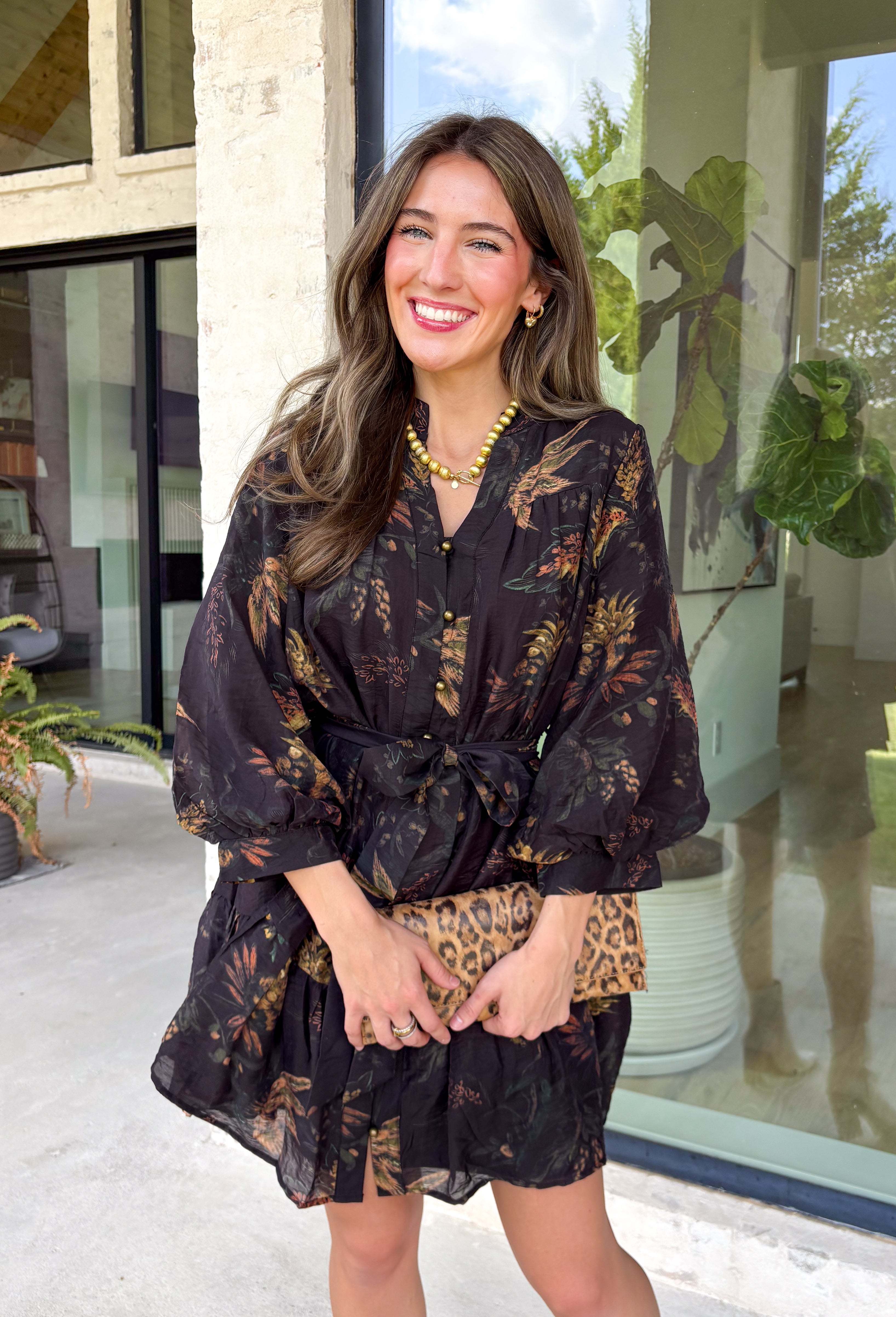 Step into the season with effortless style in this beautifully detailed dress from Entro. Designed with a classic black backdrop and a rich floral print in shades of brown and tan, this timeless dress captures the warmth of fall with every detail. The flattering v-neckline and button-front design create a polished silhouette, while the included matching belt defines the waist for a customizable fit. Finished with ¾ balloon sleeves, a playful ruffle hem, convenient side pockets.