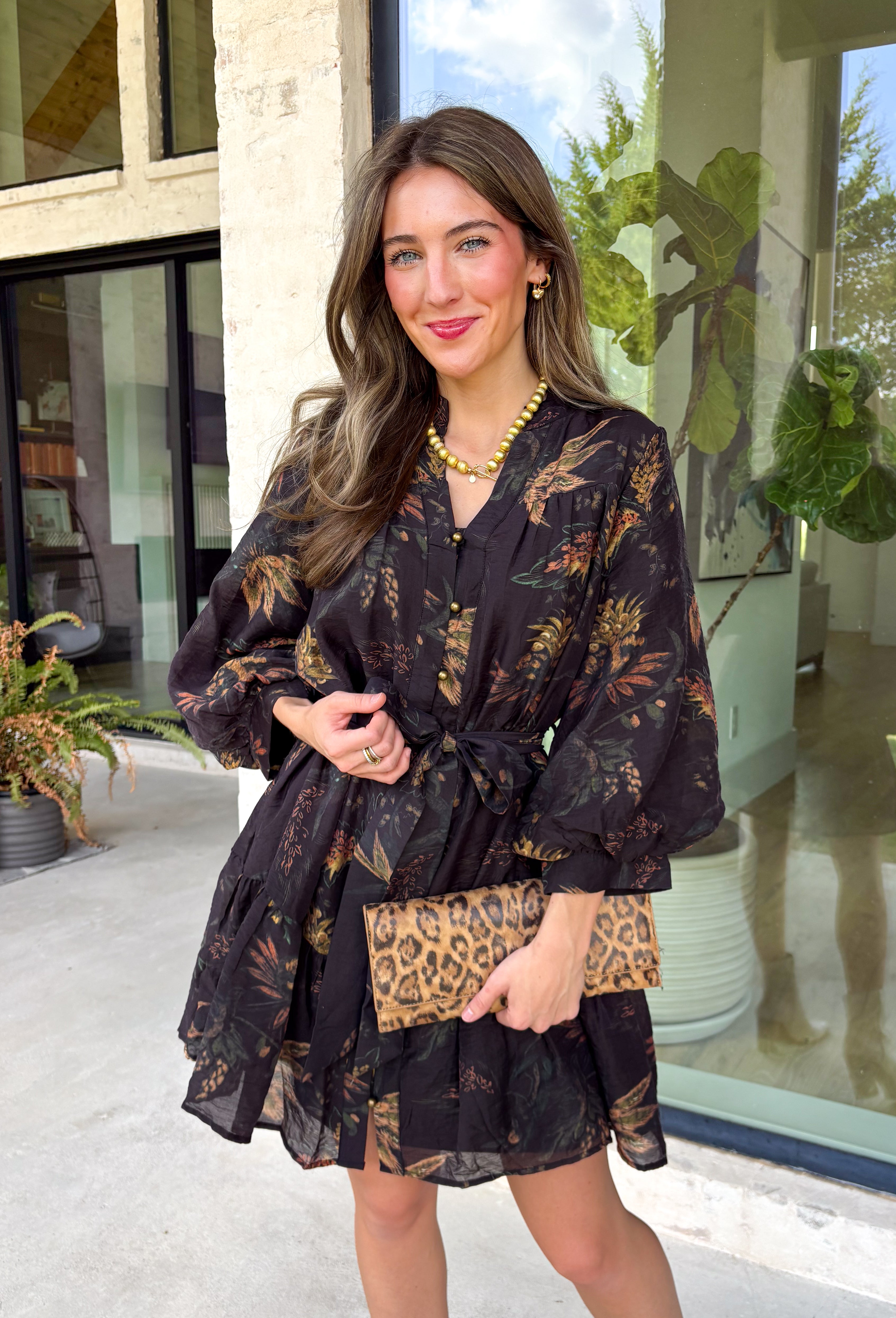 Step into the season with effortless style in this beautifully detailed dress from Entro. Designed with a classic black backdrop and a rich floral print in shades of brown and tan, this timeless dress captures the warmth of fall with every detail. The flattering v-neckline and button-front design create a polished silhouette, while the included matching belt defines the waist for a customizable fit. Finished with ¾ balloon sleeves, a playful ruffle hem, convenient side pockets.