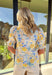 Soft, sunny, and full of spring charm, this off-white short sleeve top by Entro is an easy favorite for the season. Featuring a yellow and blue floral print, it feels bright and cheerful without being overpowering. The coordinating yellow and blue band along the front adds a fun pop of contrast, while the smocked neckline and ruffle sleeves bring a sweet, feminine finish. Crafted from breathable cotton, this top is perfect for warm spring days.