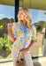 Soft, sunny, and full of spring charm, this off-white short sleeve top by Entro is an easy favorite for the season. Featuring a yellow and blue floral print, it feels bright and cheerful without being overpowering. The coordinating yellow and blue band along the front adds a fun pop of contrast, while the smocked neckline and ruffle sleeves bring a sweet, feminine finish. Crafted from breathable cotton, this top is perfect for warm spring days.