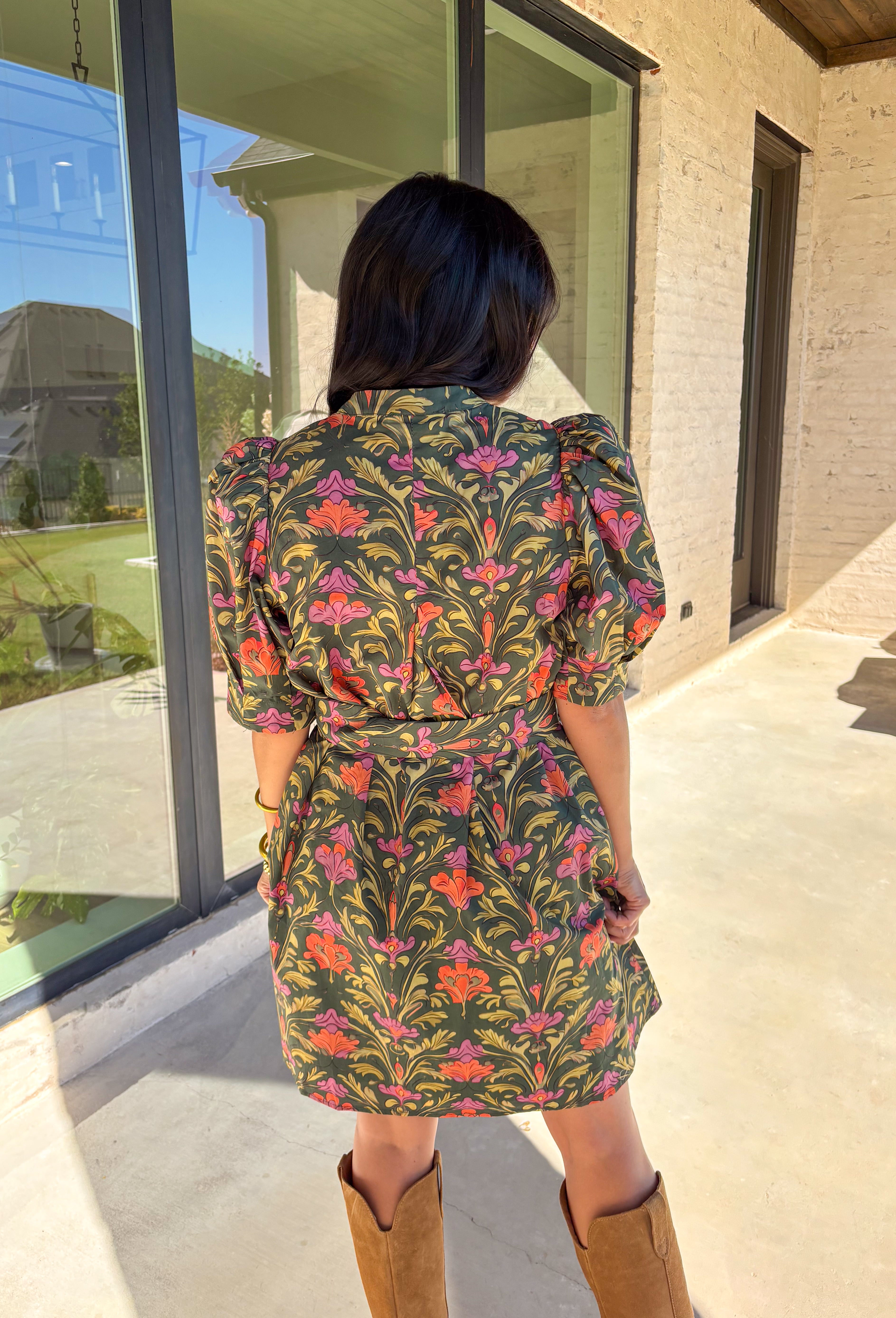 This dark green dress by Entro is a fall favorite with its vibrant botanical print and timeless silhouette. Featuring puff sleeves and pintuck detailing at the front, it brings just the right amount of texture and charm. The matching buckle belt adds shape at the waist, while the button-front closure and side pockets make it as functional as it is flattering. Designed in olive, orange, and pink tones, it’s the perfect balance of classic and playful—made for everything from brunch to fall festivals.