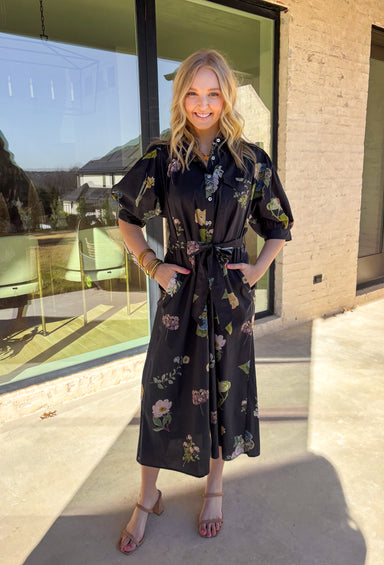Classic with a fresh spring update, this black short sleeve midi dress by Entro is an effortless go-to for the season. Featuring a soft floral print in shades of green, blue, and pink, it adds just the right amount of color to a timeless silhouette. The collared neckline, chest pocket, and button-down front give it a clean, structured look, while the matching self-tie waist creates a flattering, adjustable fit. Finished with side pockets and a breathable cotton lining, this dress is as practical as it is pr