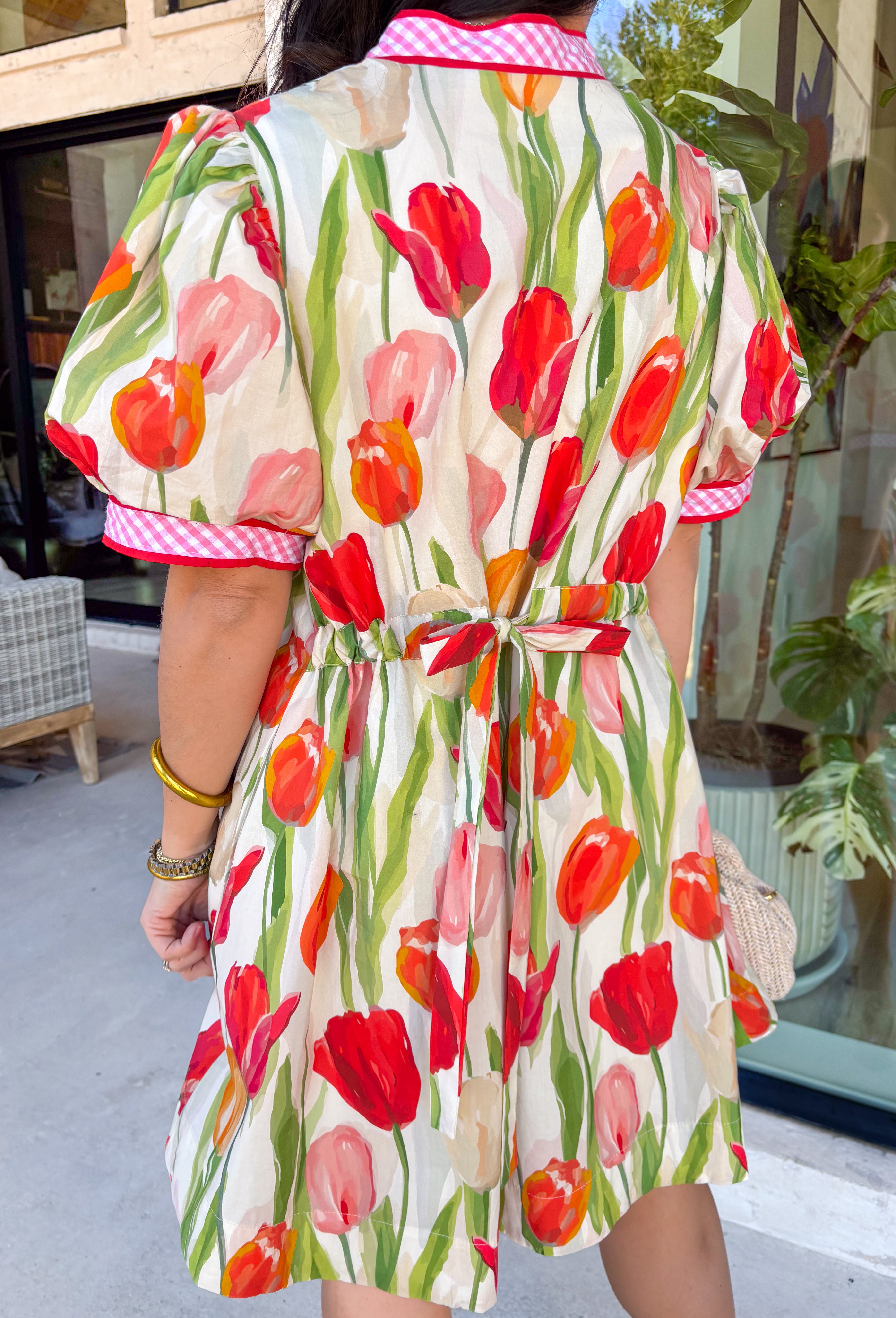 Bold blooms meet playful details in this neutral short sleeve dress that’s made to turn heads this spring. Featuring a vibrant tulip floral print in red, green, orange, and pink, it brings all the happy color while still feeling easy and wearable. The front button detail is accented with red trim and a pink-and-white gingham print for an extra pop of charm. Puff sleeves add a feminine touch, while the adjustable tie detail in the back creates a flattering, customizable fit. 