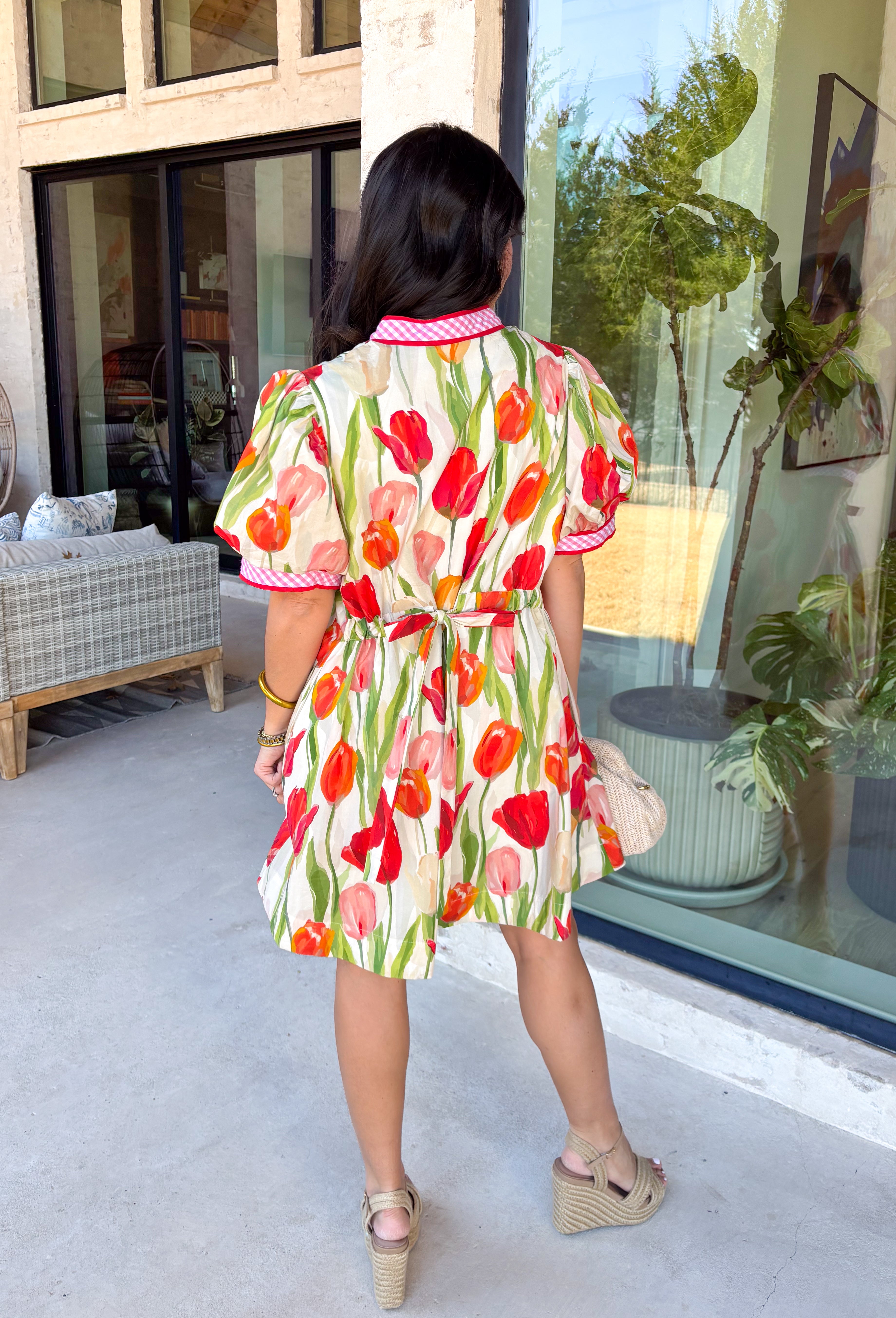 Bold blooms meet playful details in this neutral short sleeve dress that’s made to turn heads this spring. Featuring a vibrant tulip floral print in red, green, orange, and pink, it brings all the happy color while still feeling easy and wearable. The front button detail is accented with red trim and a pink-and-white gingham print for an extra pop of charm. Puff sleeves add a feminine touch, while the adjustable tie detail in the back creates a flattering, customizable fit. 