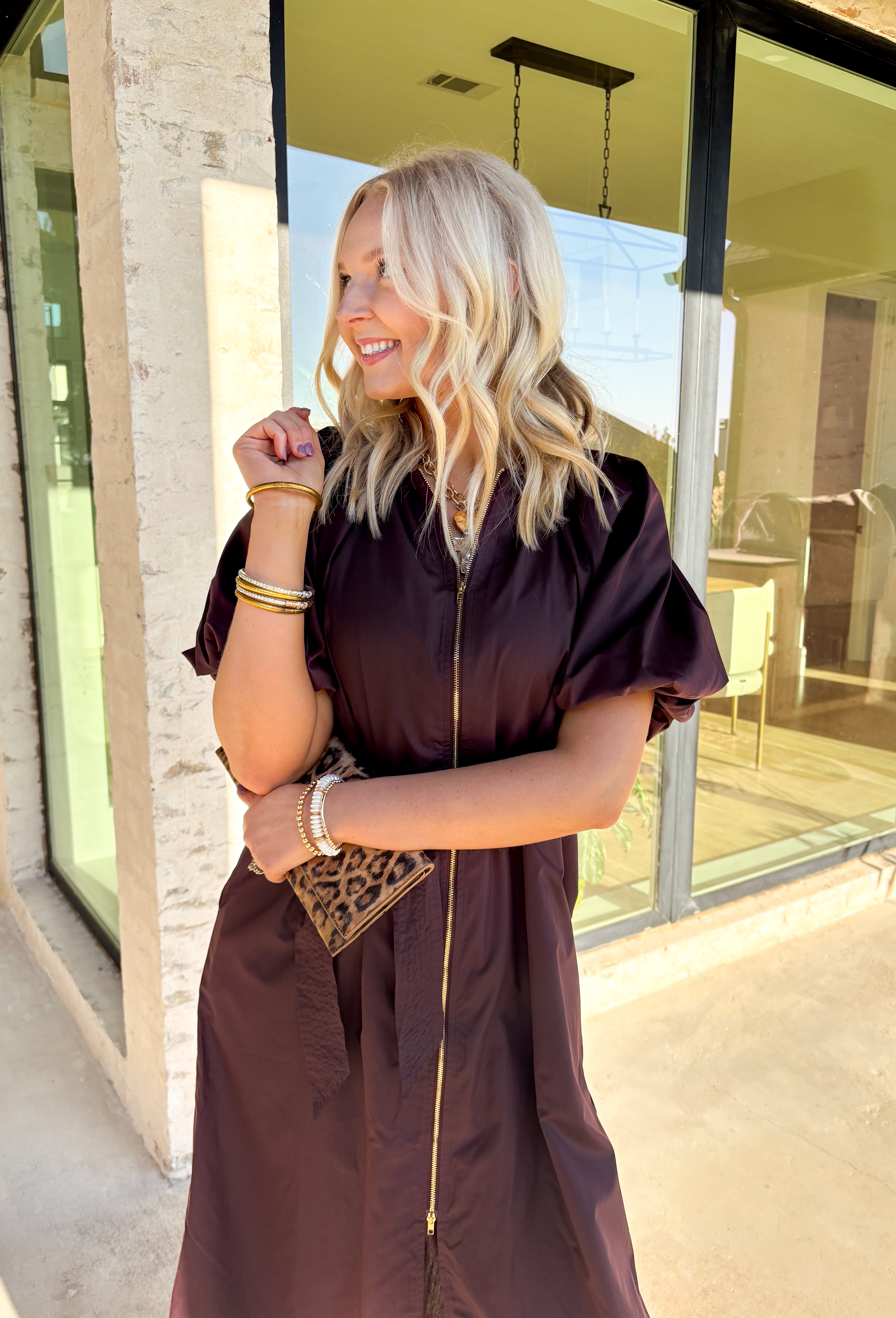 This chocolate brown midi dress by Entro is a flattering mix of modern detail and timeless design. The short puff sleeves and gathered neckline give it a feminine touch, while the exposed front zipper adds a hint of edge. A removable self-tie belt lets you define your waist, and side pockets keep it practical for everyday wear. Fully lined and easy to style, this dress is perfect for transitioning from casual afternoons to evening plans.