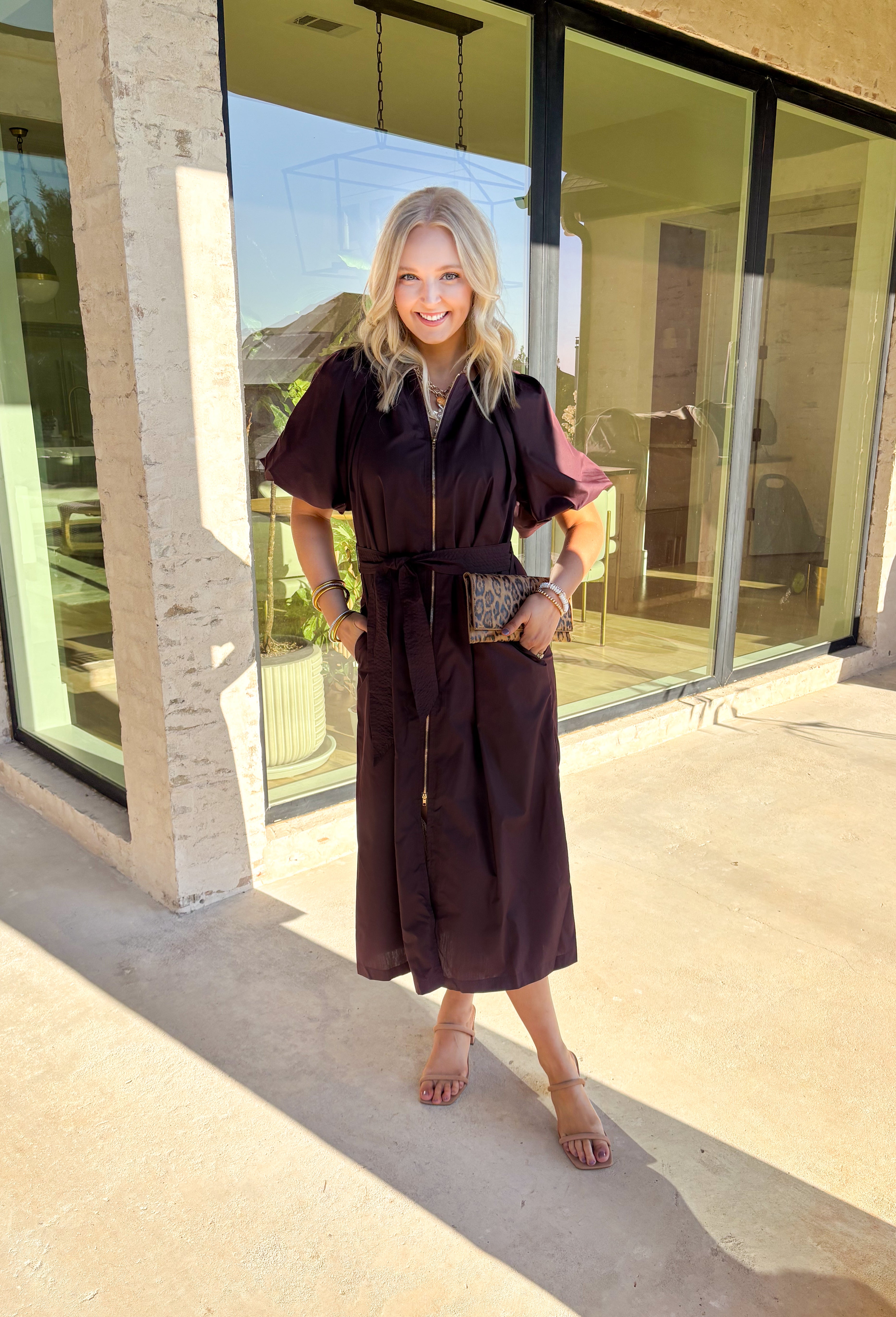 This chocolate brown midi dress by Entro is a flattering mix of modern detail and timeless design. The short puff sleeves and gathered neckline give it a feminine touch, while the exposed front zipper adds a hint of edge. A removable self-tie belt lets you define your waist, and side pockets keep it practical for everyday wear. Fully lined and easy to style, this dress is perfect for transitioning from casual afternoons to evening plans.