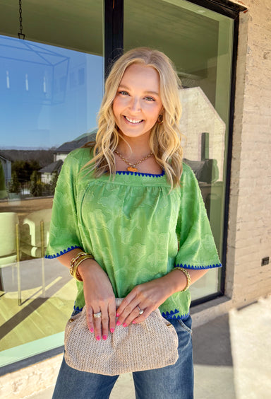 Fresh, fun, and full of texture, this green short sleeve top by Voy is an easy spring standout. Designed with a monochromatic textured floral print, it adds dimension and detail while keeping the look clean and wearable. Royal blue embroidered trim along the front, cuffs, and hem gives it a bold pop of contrast, and the square neckline adds a flattering, modern finish. Lightweight and easy to style, this top is perfect for brightening up your everyday spring outfits.