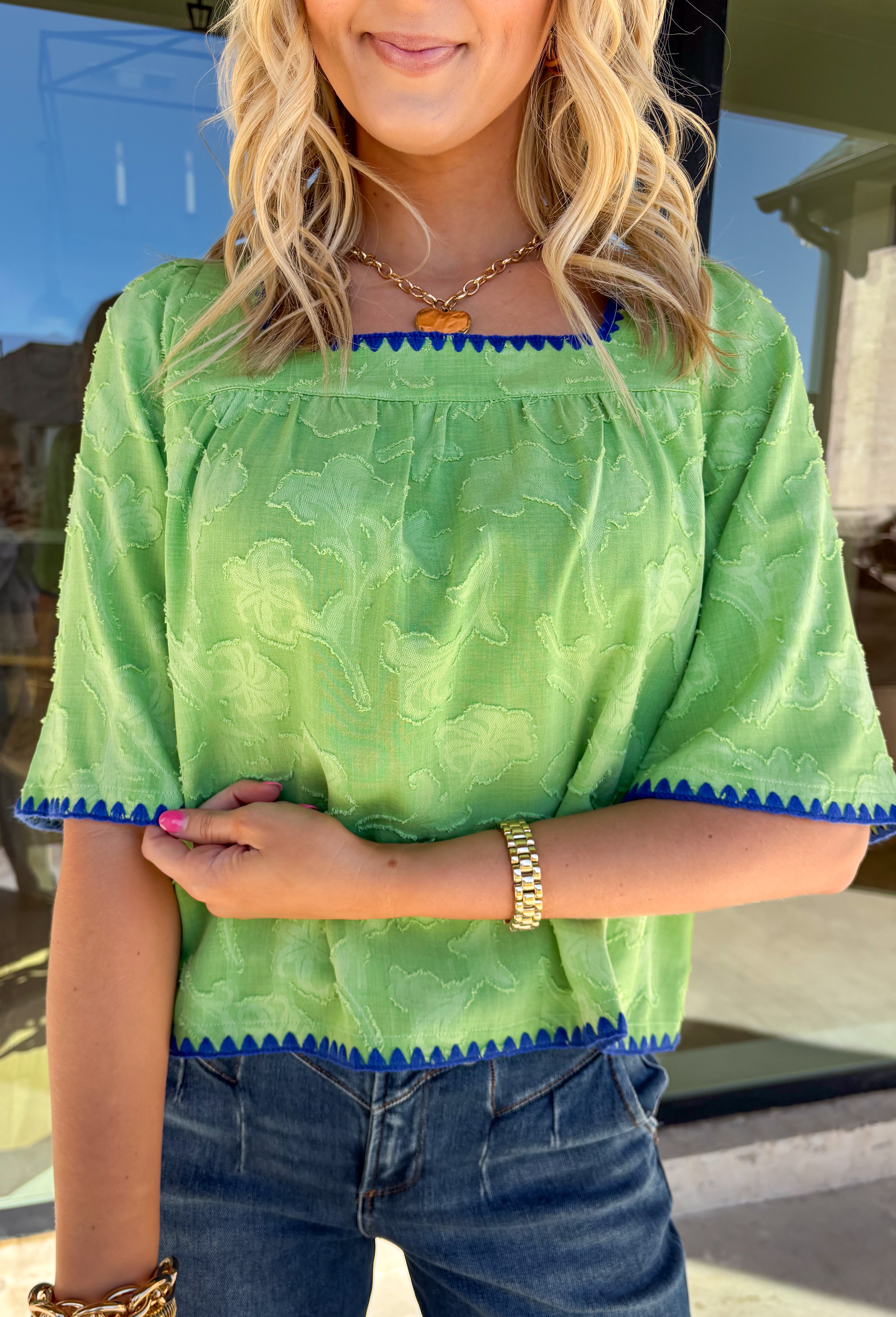 Fresh, fun, and full of texture, this green short sleeve top by Voy is an easy spring standout. Designed with a monochromatic textured floral print, it adds dimension and detail while keeping the look clean and wearable. Royal blue embroidered trim along the front, cuffs, and hem gives it a bold pop of contrast, and the square neckline adds a flattering, modern finish. Lightweight and easy to style, this top is perfect for brightening up your everyday spring outfits.