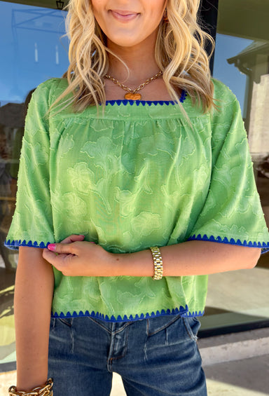 Fresh, fun, and full of texture, this green short sleeve top by Voy is an easy spring standout. Designed with a monochromatic textured floral print, it adds dimension and detail while keeping the look clean and wearable. Royal blue embroidered trim along the front, cuffs, and hem gives it a bold pop of contrast, and the square neckline adds a flattering, modern finish. Lightweight and easy to style, this top is perfect for brightening up your everyday spring outfits.