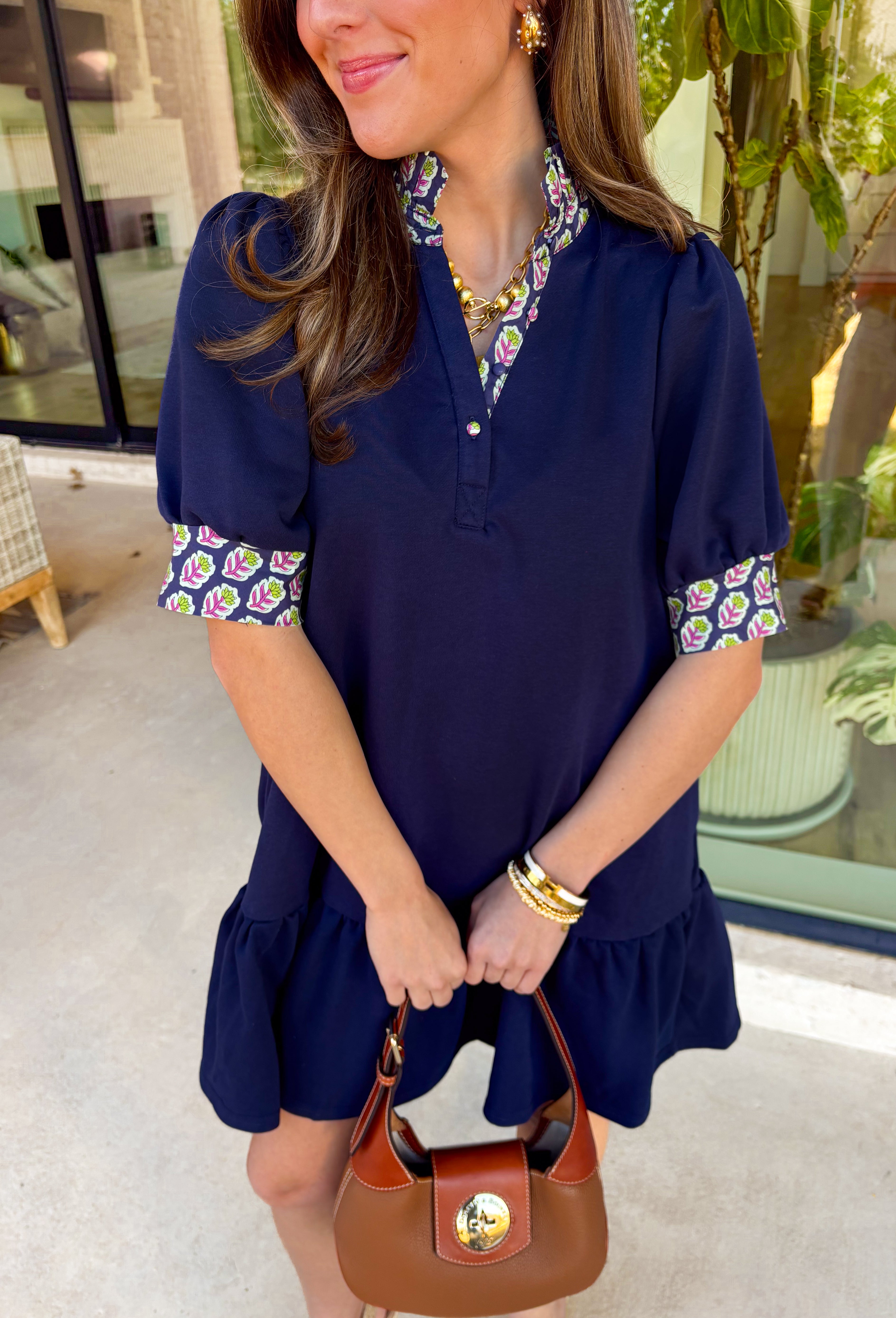 This navy dress is the definition of easy spring style with a little something special! The soft tiered skirt gives it that breezy, swishy movement we love, while the purple and green floral print along the neckline and sleeve cuffs adds the prettiest pop of color. Finished with a single matching button detail on the front (so cute and subtle), plus side pockets for extra everyday ease—this one is made for sunny days, casual plans, and feeling effortlessly put-together.