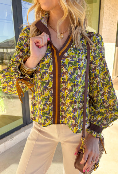 Fresh and full of spring charm, this green floral print top by &amp;Merci is an easy way to brighten your everyday look. Featuring a mix of green, blue, brown, and cream florals, this top feels both earthy and light for the season. The smocked ruffle neckline and smocked sleeves add a sweet, feminine touch, while the brown and tan trim down the front gives it a little extra detail that makes it feel special.