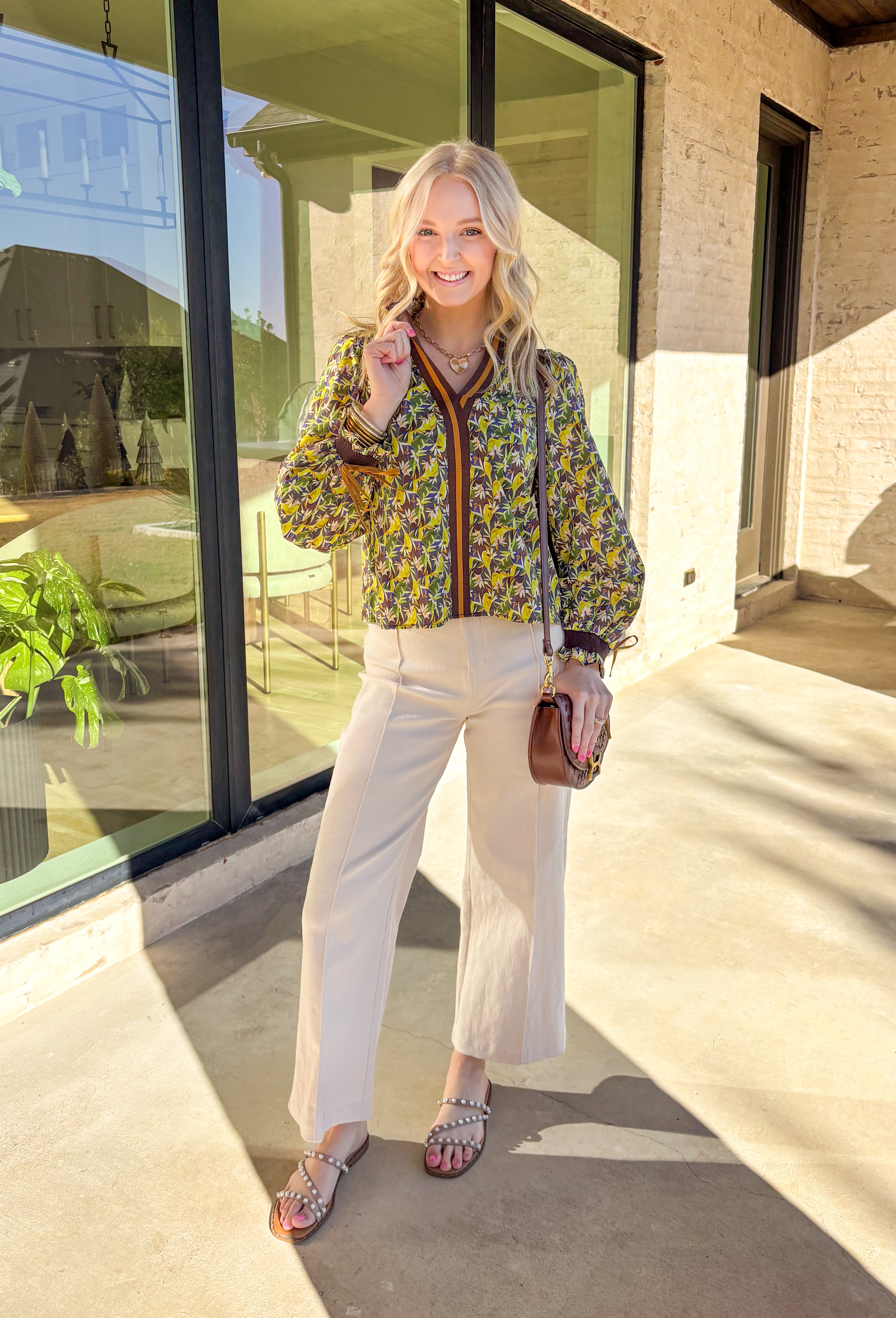 Fresh and full of spring charm, this green floral print top by &amp;Merci is an easy way to brighten your everyday look. Featuring a mix of green, blue, brown, and cream florals, this top feels both earthy and light for the season. The smocked ruffle neckline and smocked sleeves add a sweet, feminine touch, while the brown and tan trim down the front gives it a little extra detail that makes it feel special.