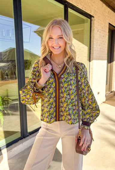 Fresh and full of spring charm, this green floral print top by &amp;Merci is an easy way to brighten your everyday look. Featuring a mix of green, blue, brown, and cream florals, this top feels both earthy and light for the season. The smocked ruffle neckline and smocked sleeves add a sweet, feminine touch, while the brown and tan trim down the front gives it a little extra detail that makes it feel special.