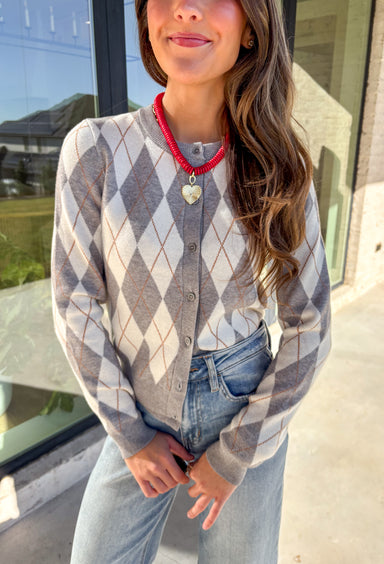 Classic pattern, cozy feel—this button-down cardigan by Haute Monde is the perfect layering piece for the season. Featuring a timeless gray and off-white argyle print with tan accents, it’s a modern twist on a preppy staple. The button-front design and soft knit fabric make it an easy go-to whether you’re dressing up for work or keeping it casual on weekends.