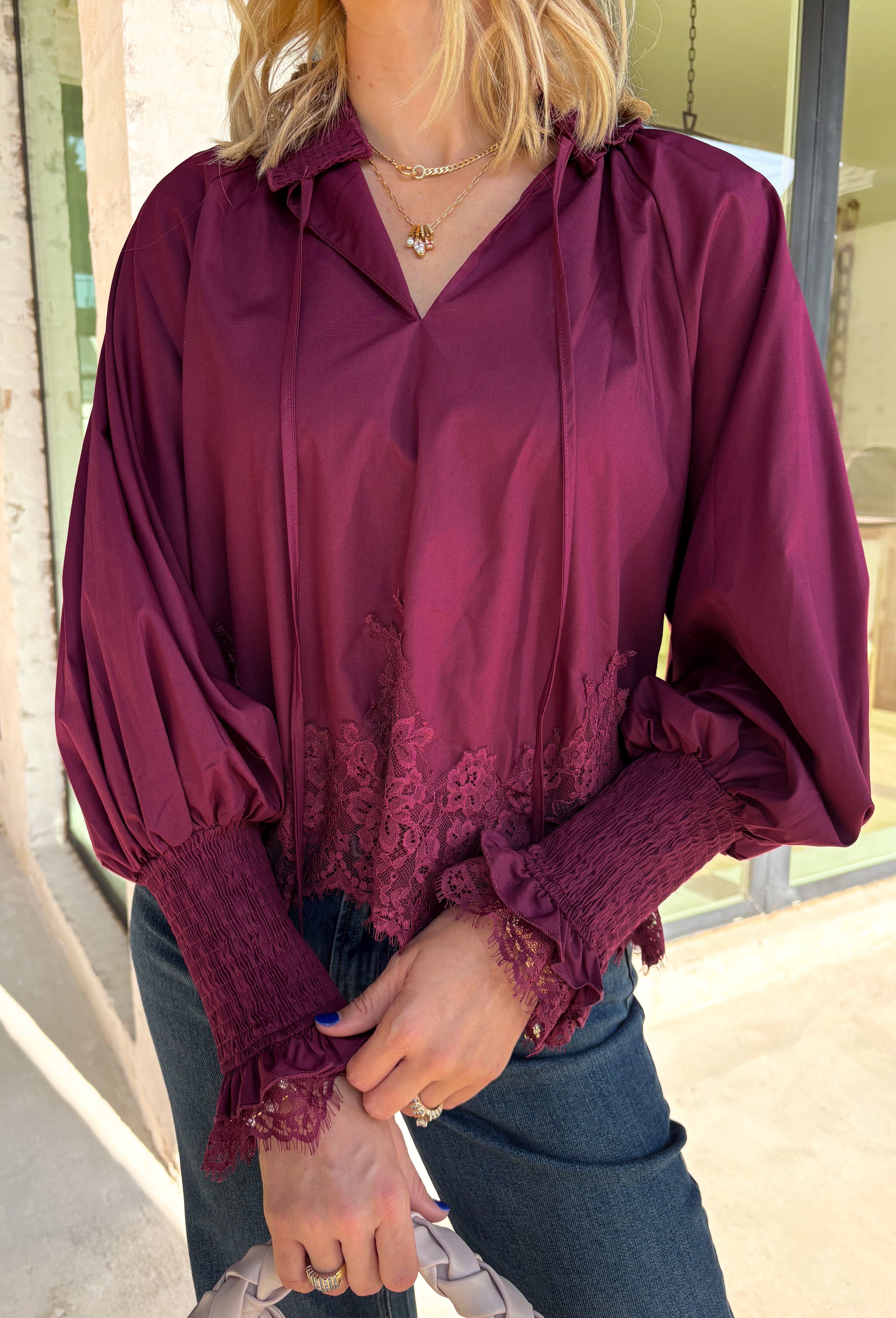 Bring a touch of romantic charm to your fall wardrobe with this beautifully detailed blouse from Entro. Designed in a rich burgundy hue, this feminine top features a softly smocked neckline with a delicate front tie for an effortlessly graceful look. The long sleeves are finished with smocked cuffs and lace trim, while the lace hem adds an elegant finishing touch. 