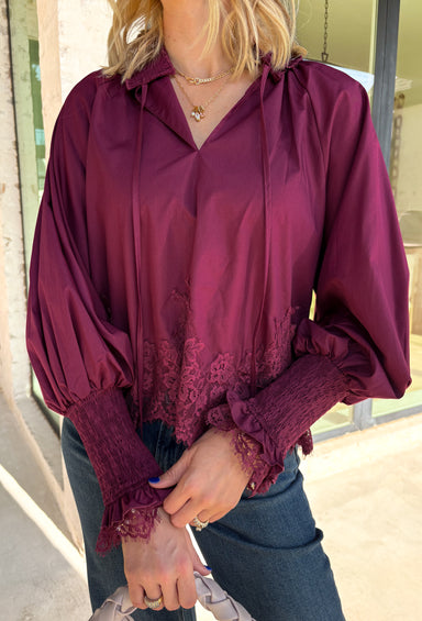 Bring a touch of romantic charm to your fall wardrobe with this beautifully detailed blouse from Entro. Designed in a rich burgundy hue, this feminine top features a softly smocked neckline with a delicate front tie for an effortlessly graceful look. The long sleeves are finished with smocked cuffs and lace trim, while the lace hem adds an elegant finishing touch. 