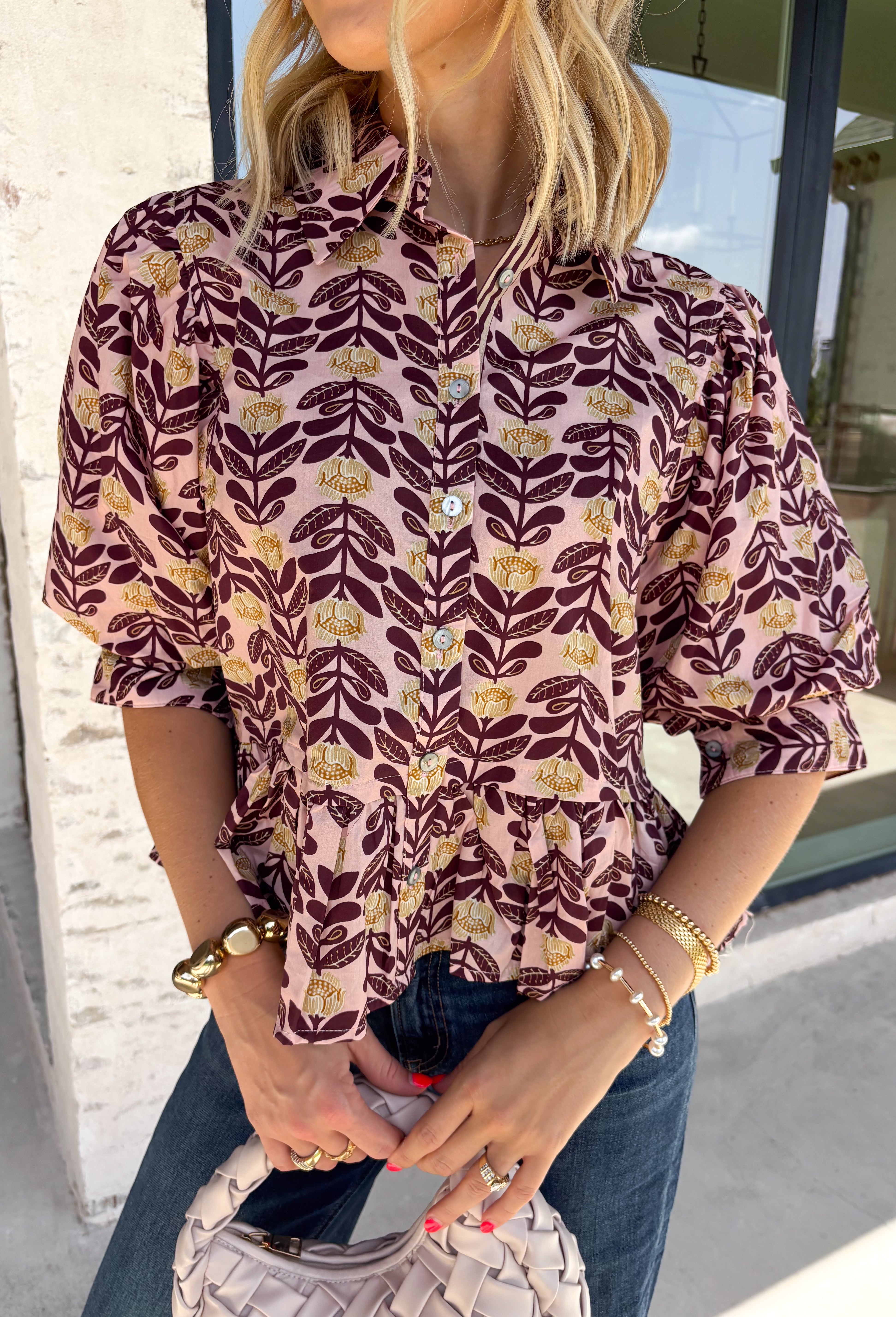 Soft, feminine, and full of seasonal charm, this floral blouse is an effortless addition to your fall wardrobe. Designed with a beautiful blush backdrop and a rich burgundy and tan floral print, this top brings together warm autumn hues in a timeless silhouette. The button-front design adds classic appeal, while the ruffle hem creates a flattering, feminine finish. Gathered detailing at the back yoke gives this blouse a relaxed, flowy fit that's perfect for everything from workdays to weekend outings.