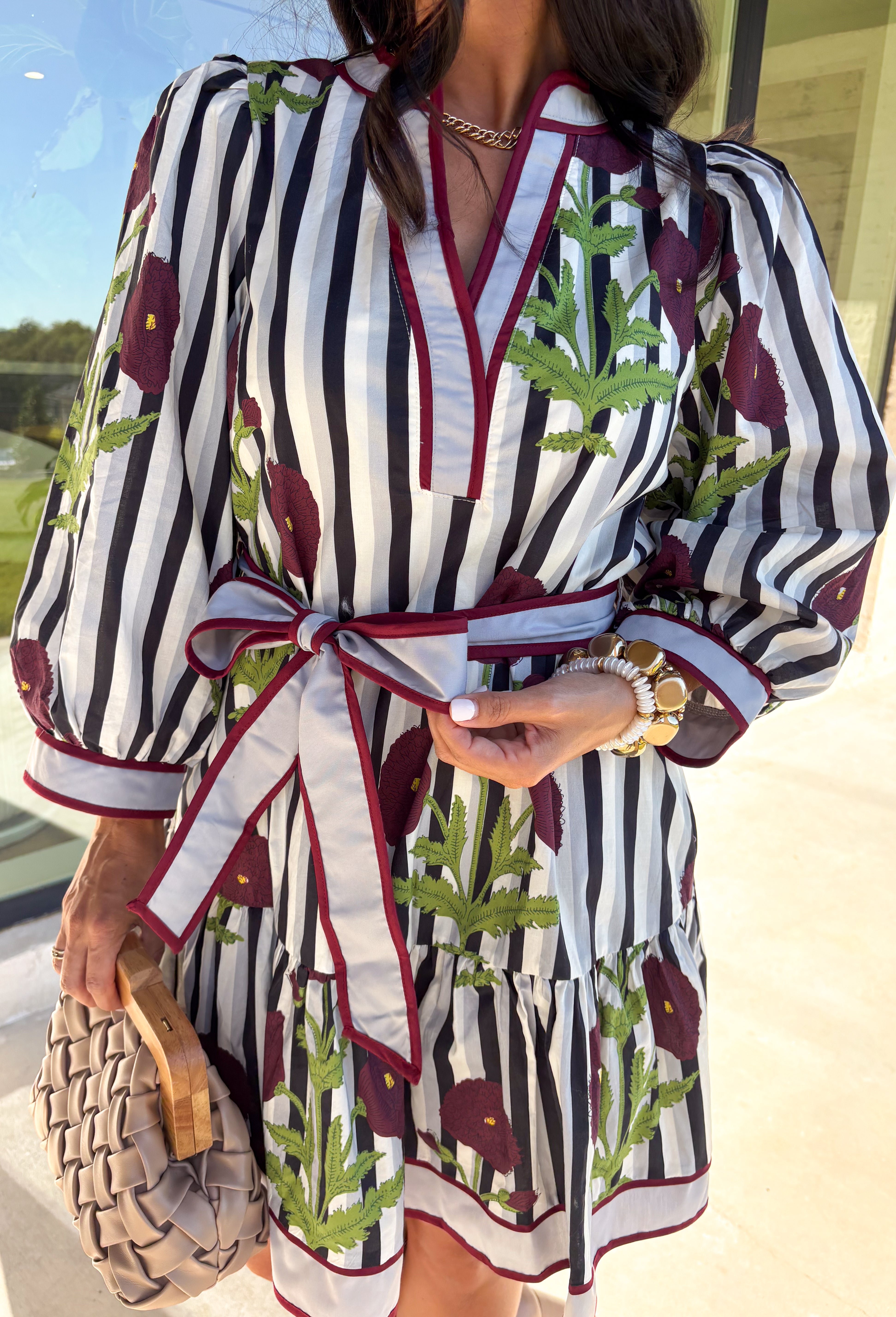 This floral stripe mini dress by Entro blends playful charm with seasonal flair. Featuring a gray, navy, light blue, off white and&nbsp; stripe base adorned with burgundy florals and green leaves, it’s the perfect balance of classic and bold. The ¾ puff sleeves add volume, while the v-neckline with contrast trim and adjustable self-tie belt create a flattering silhouette. A ruffle hem and side pockets finish the look, with a full cotton lining that makes it as practical as it is pretty.