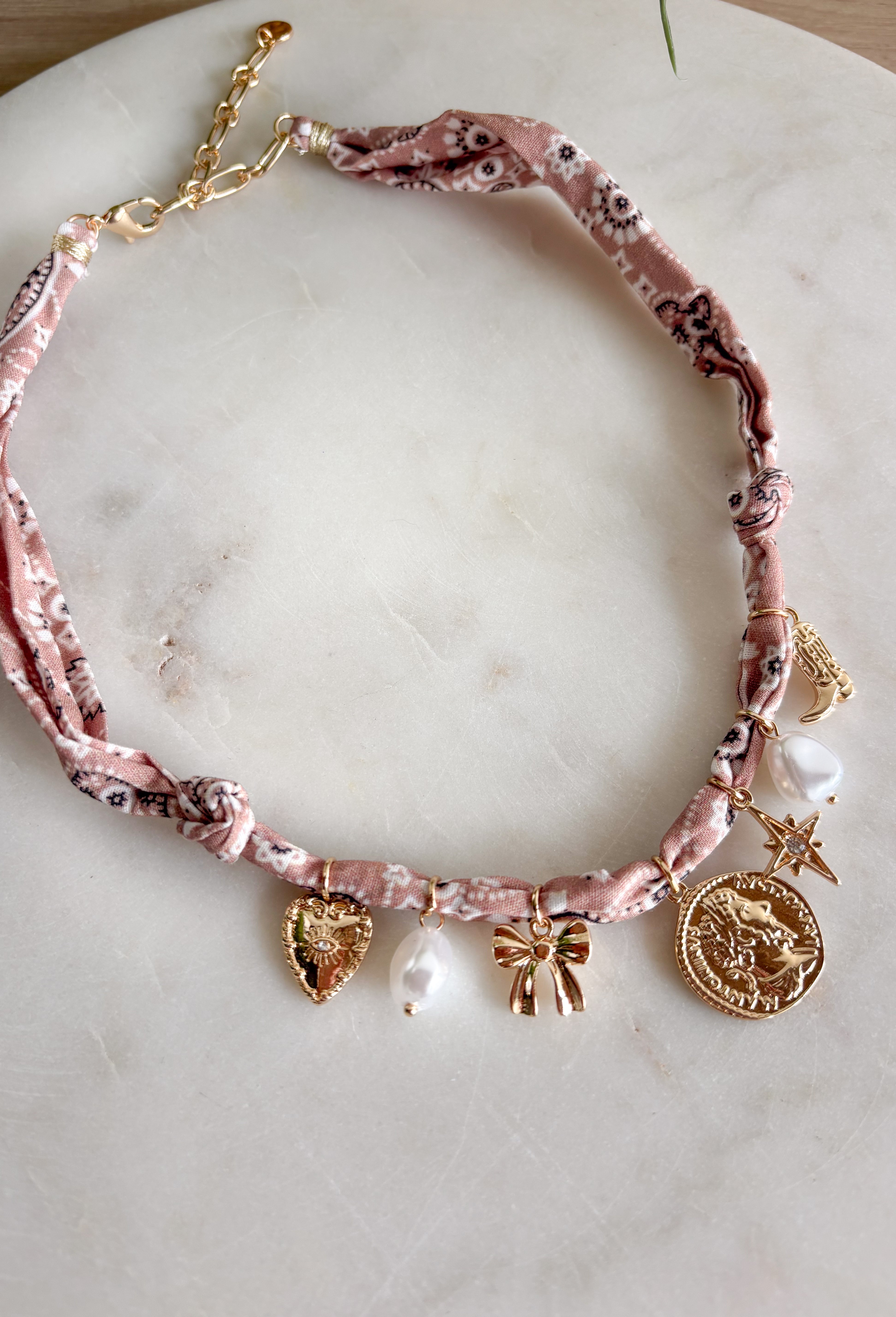 Playful and trendy, this tan bandana necklace is full of personality. Detailed with a mix of eclectic charms—including a sparkling crystal star, gold bow, pearls, gold pendant, gold heart, and a gold cowboy boot—it’s the perfect statement piece to add fun flair to any outfit. With a 16" length and 3" extender, you can adjust it to layer with other necklaces or let it shine on its own. A must-have for adding a pop of charm to your wardrobe.