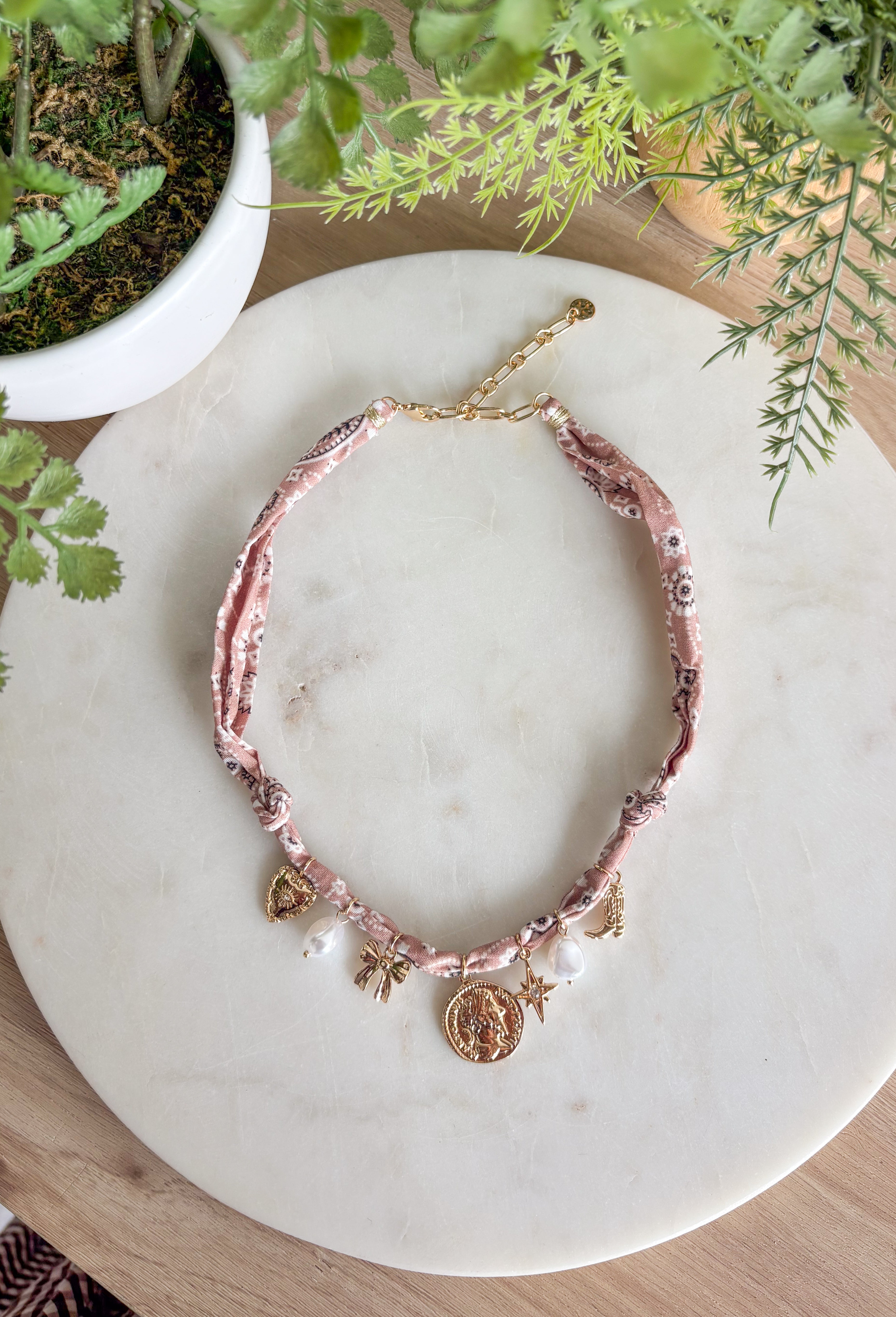 Playful and trendy, this tan bandana necklace is full of personality. Detailed with a mix of eclectic charms—including a sparkling crystal star, gold bow, pearls, gold pendant, gold heart, and a gold cowboy boot—it’s the perfect statement piece to add fun flair to any outfit. With a 16" length and 3" extender, you can adjust it to layer with other necklaces or let it shine on its own. A must-have for adding a pop of charm to your wardrobe.
