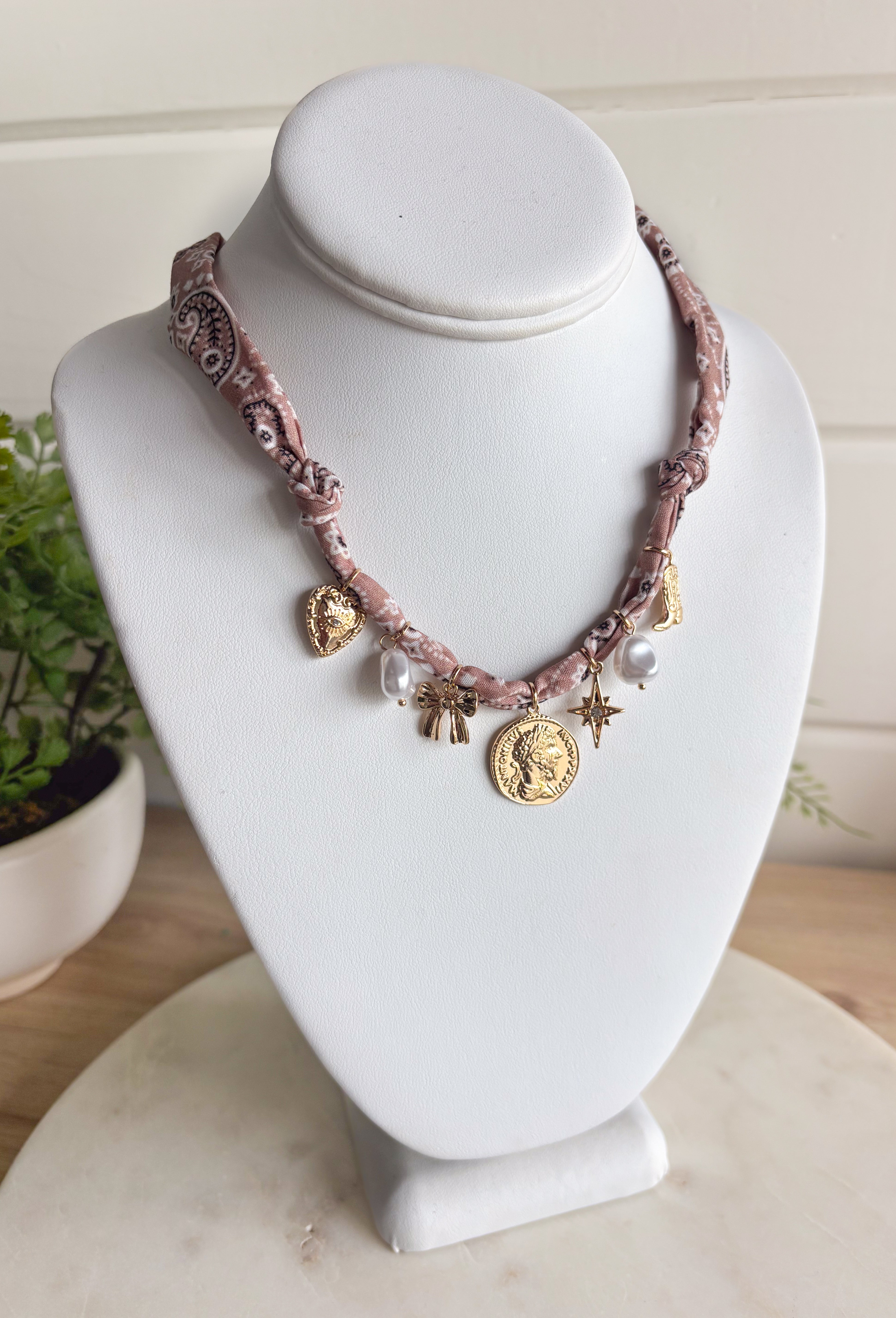 Playful and trendy, this tan bandana necklace is full of personality. Detailed with a mix of eclectic charms—including a sparkling crystal star, gold bow, pearls, gold pendant, gold heart, and a gold cowboy boot—it’s the perfect statement piece to add fun flair to any outfit. With a 16" length and 3" extender, you can adjust it to layer with other necklaces or let it shine on its own. A must-have for adding a pop of charm to your wardrobe.