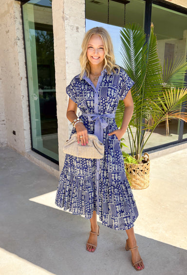 Fresh, charming, and full of thoughtful details, this blue button-down midi dress by Entro feels like spring in dress form. Featuring an off-white floral print with delicate mini star accents throughout, it strikes the perfect balance between playful and classic. The blue and white striped collar and trim add a crisp contrast, while the matching self-tie waist creates a flattering, easy fit. 