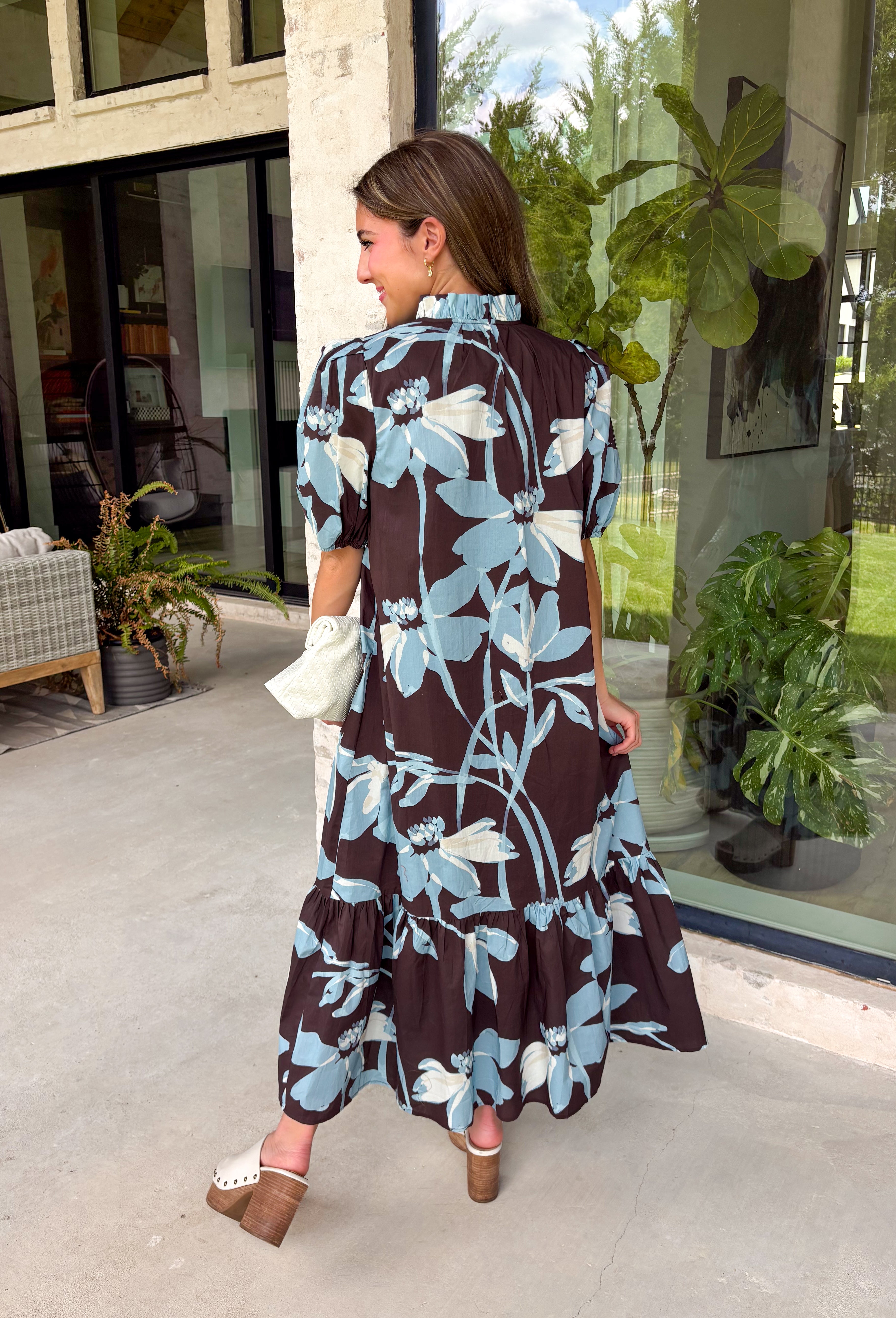 Celebrate the beauty of the season in this effortlessly feminine floral midi dress. Featuring a rich brown backdrop with a bold light blue and white floral print, this timeless silhouette is perfect for everything from weekend brunches to holiday gatherings. The ruffle neckline, puff sleeves, and flowy ruffle hem add a soft, romantic touch, while the button-front design creates a classic look you'll wear year after year. 