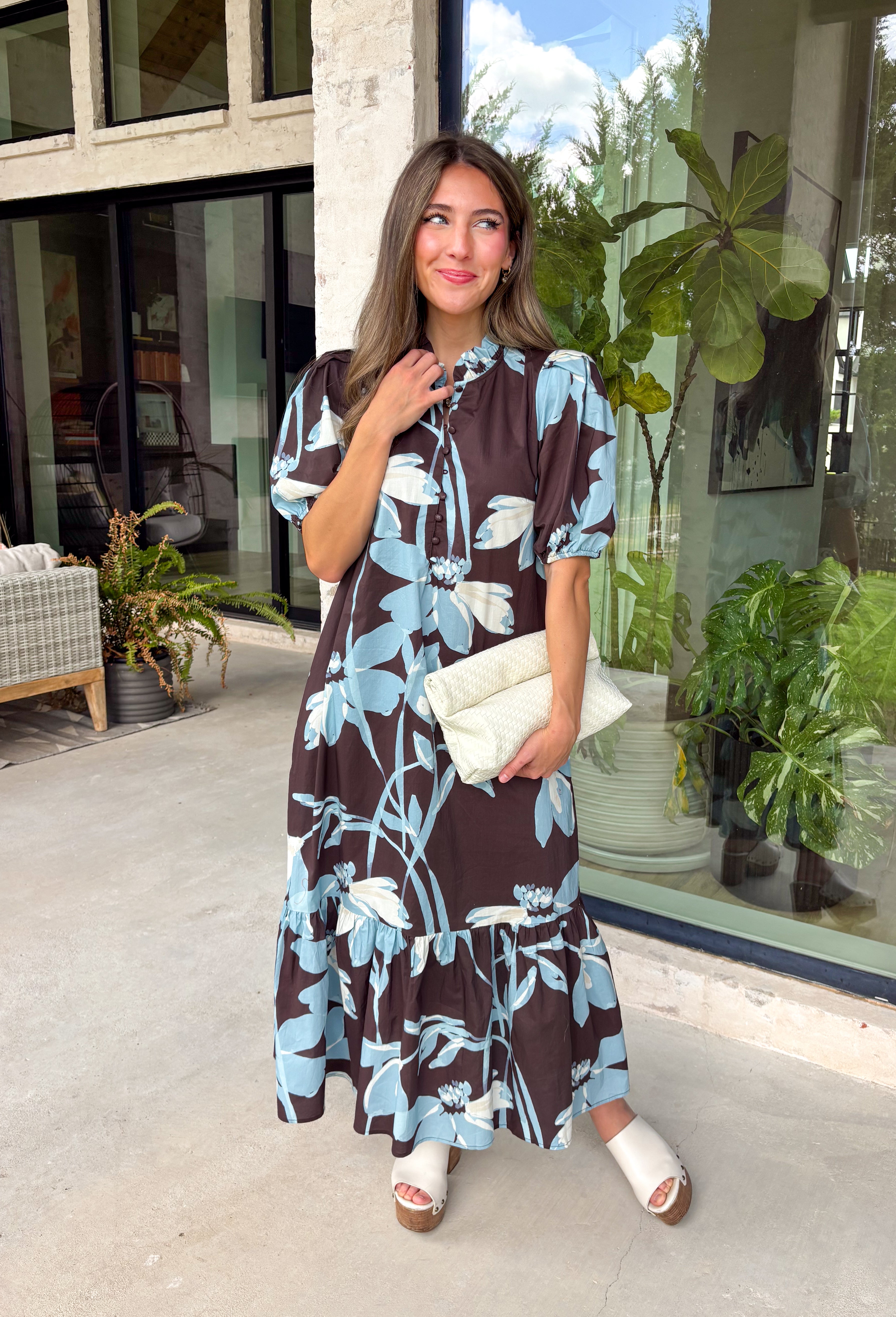 Celebrate the beauty of the season in this effortlessly feminine floral midi dress. Featuring a rich brown backdrop with a bold light blue and white floral print, this timeless silhouette is perfect for everything from weekend brunches to holiday gatherings. The ruffle neckline, puff sleeves, and flowy ruffle hem add a soft, romantic touch, while the button-front design creates a classic look you'll wear year after year. 