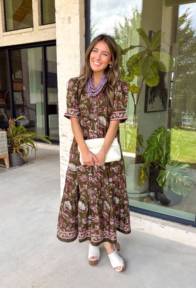 Bring vibrant seasonal color to your wardrobe with this beautifully feminine midi dress from Entro. Designed with a rich olive backdrop and a stunning floral print in shades of magenta and white, this timeless dress is perfect for everything from brunch dates to family gatherings. The button-front design and subtle ruffle neckline create a classic, polished look, while the puff sleeves add just the right touch of charm.