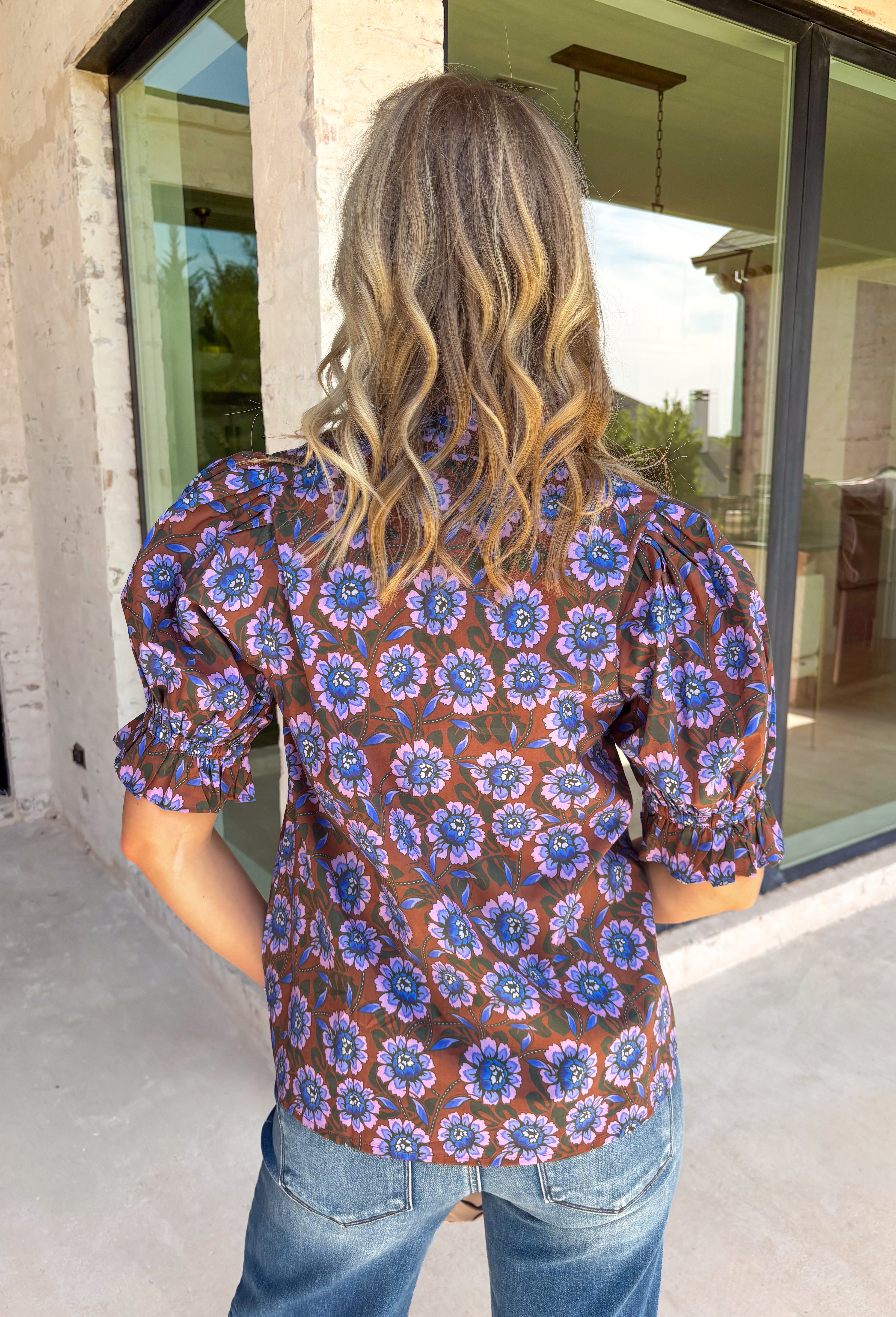 Fresh florals meet rich autumn colors in this beautifully feminine top. Featuring a warm brown backdrop with blooms in shades of blue, purple, and green, this top is an effortless way to brighten your fall wardrobe. The ruffled accent neckline and flattering v-neck add timeless charm, while the short puff sleeves with elastic ruffle cuffs create a soft, romantic silhouette. 
