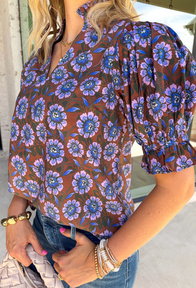 Fresh florals meet rich autumn colors in this beautifully feminine top. Featuring a warm brown backdrop with blooms in shades of blue, purple, and green, this top is an effortless way to brighten your fall wardrobe. The ruffled accent neckline and flattering v-neck add timeless charm, while the short puff sleeves with elastic ruffle cuffs create a soft, romantic silhouette. 