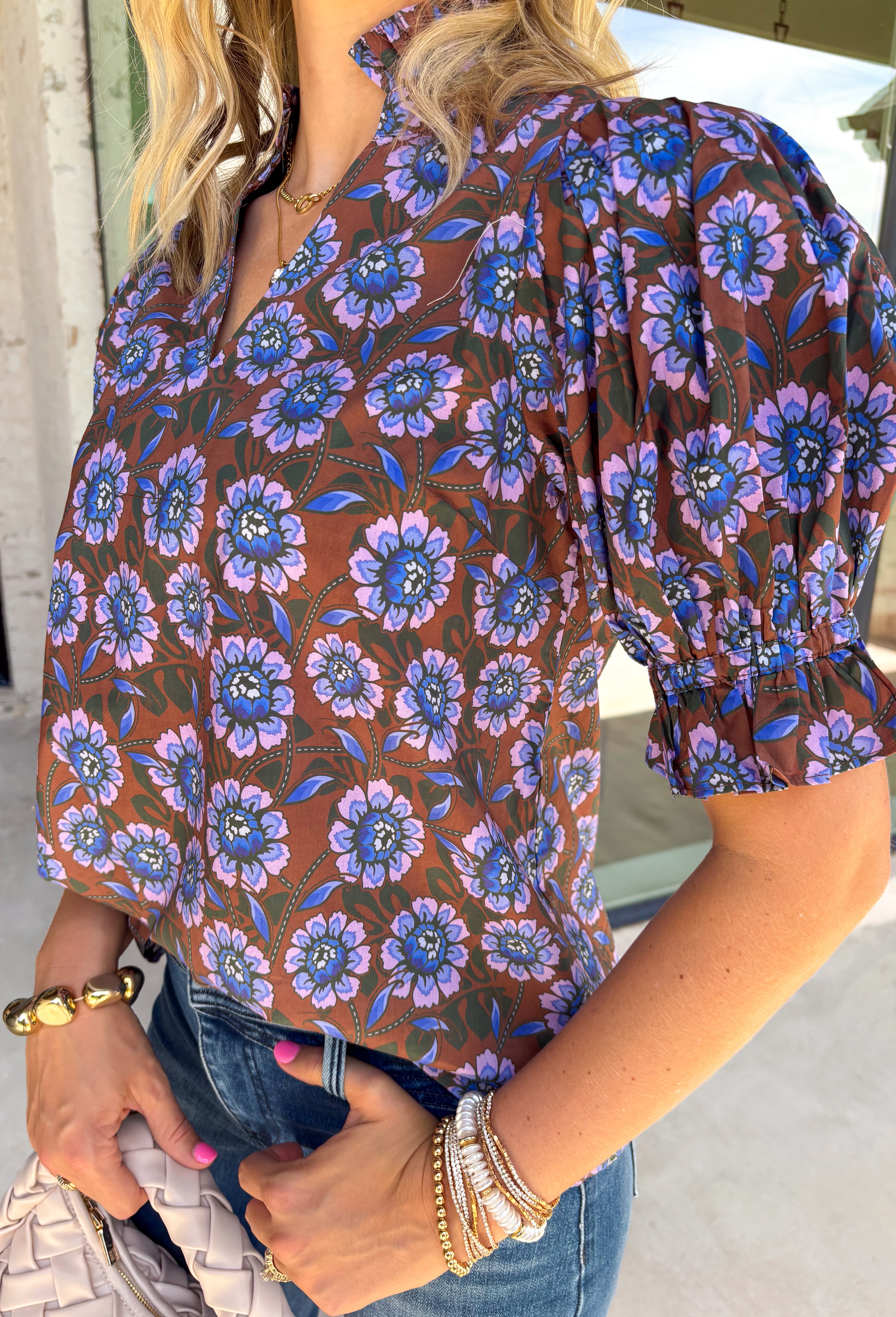 Fresh florals meet rich autumn colors in this beautifully feminine top. Featuring a warm brown backdrop with blooms in shades of blue, purple, and green, this top is an effortless way to brighten your fall wardrobe. The ruffled accent neckline and flattering v-neck add timeless charm, while the short puff sleeves with elastic ruffle cuffs create a soft, romantic silhouette. 