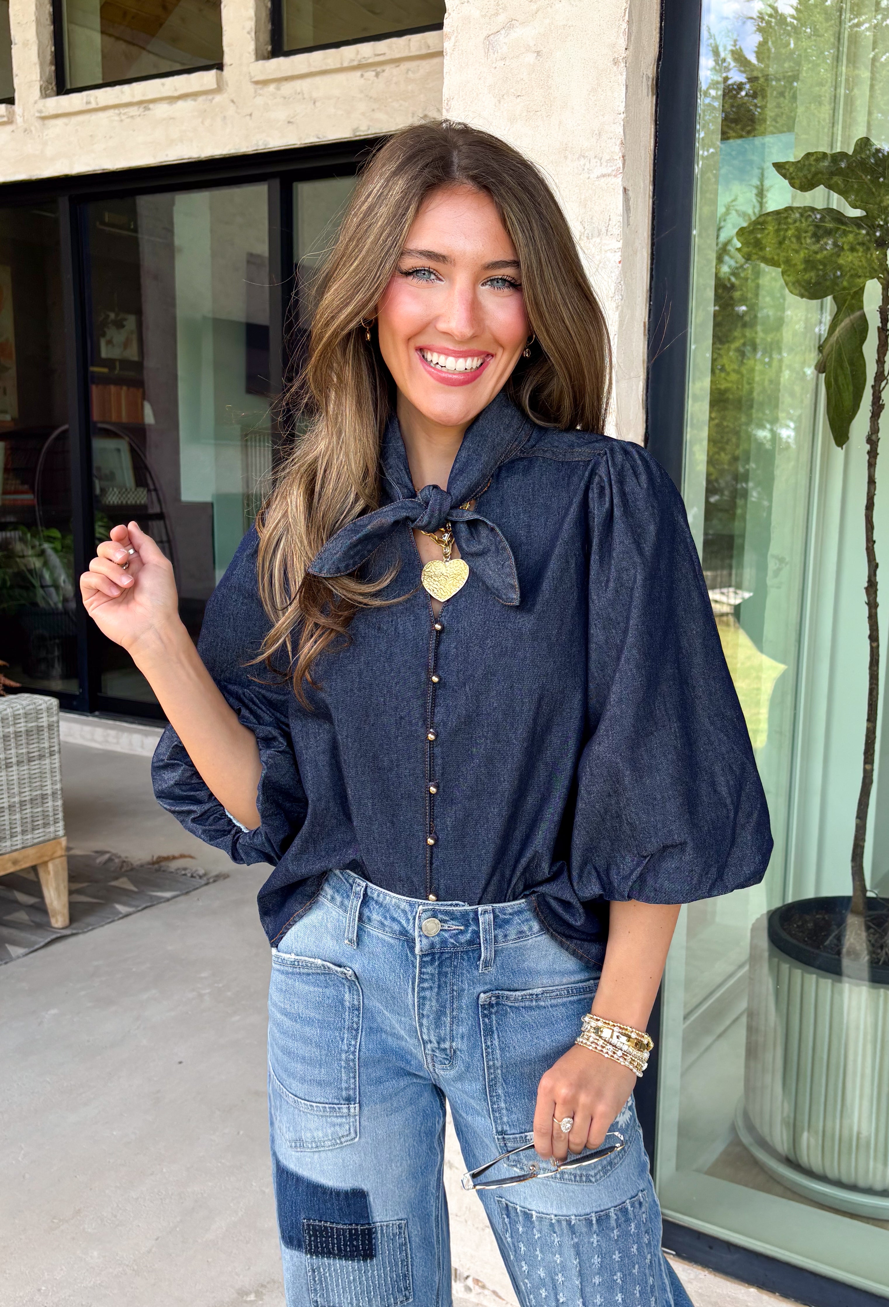 Give your denim collection a fresh twist with this dark wash blouse from Entro. The lightweight cotton fabric keeps it comfortable and easy to wear, while the rich dark wash feels right at home as fall transitions into winter. A high neckline with a self-tie detail adds a pretty focal point, complemented by gold button detailing along the front. The ¾ puff sleeves bring a little volume to the silhouette without making it feel overly dressy. 