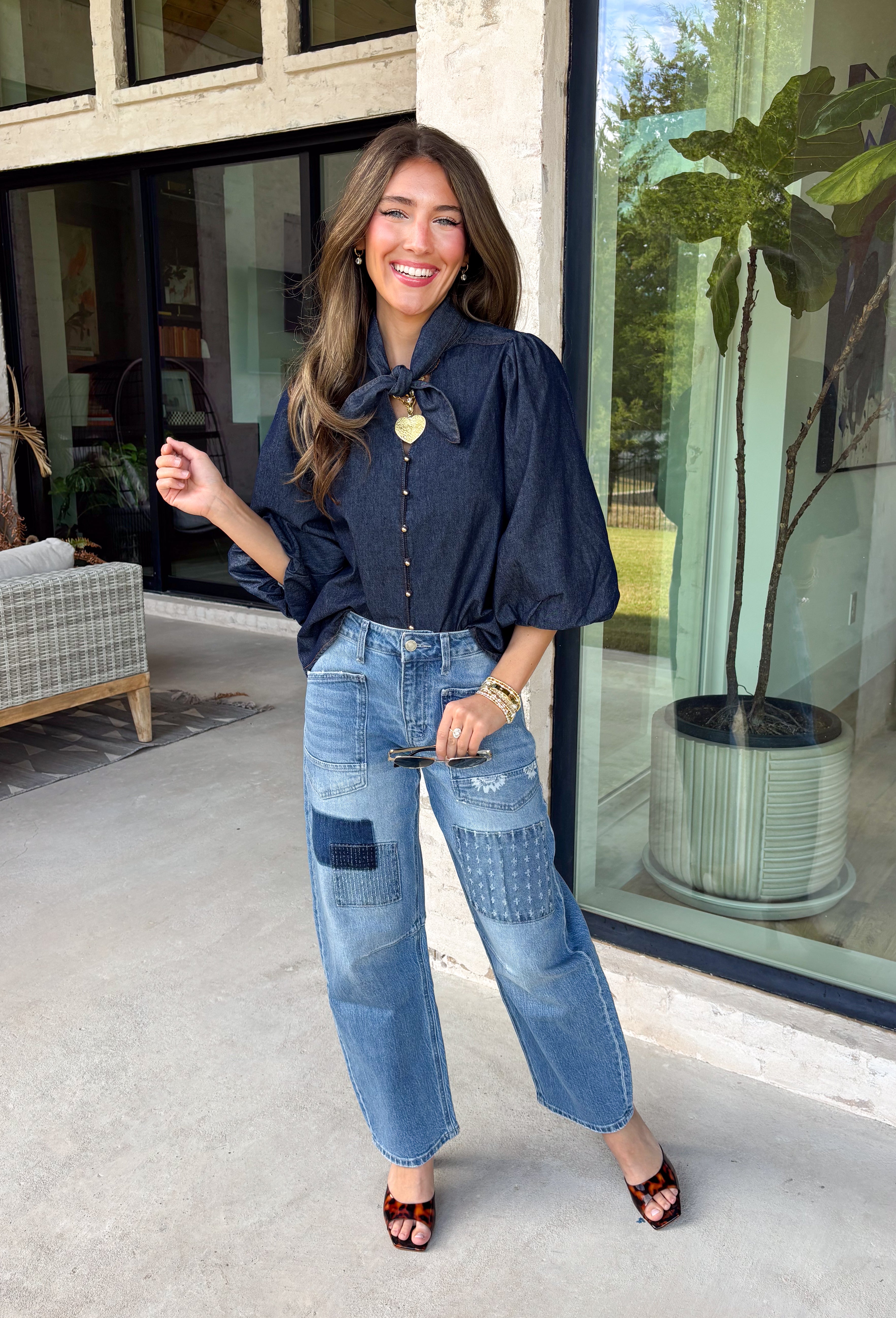 Give your denim collection a fresh twist with this dark wash blouse from Entro. The lightweight cotton fabric keeps it comfortable and easy to wear, while the rich dark wash feels right at home as fall transitions into winter. A high neckline with a self-tie detail adds a pretty focal point, complemented by gold button detailing along the front. The ¾ puff sleeves bring a little volume to the silhouette without making it feel overly dressy. 