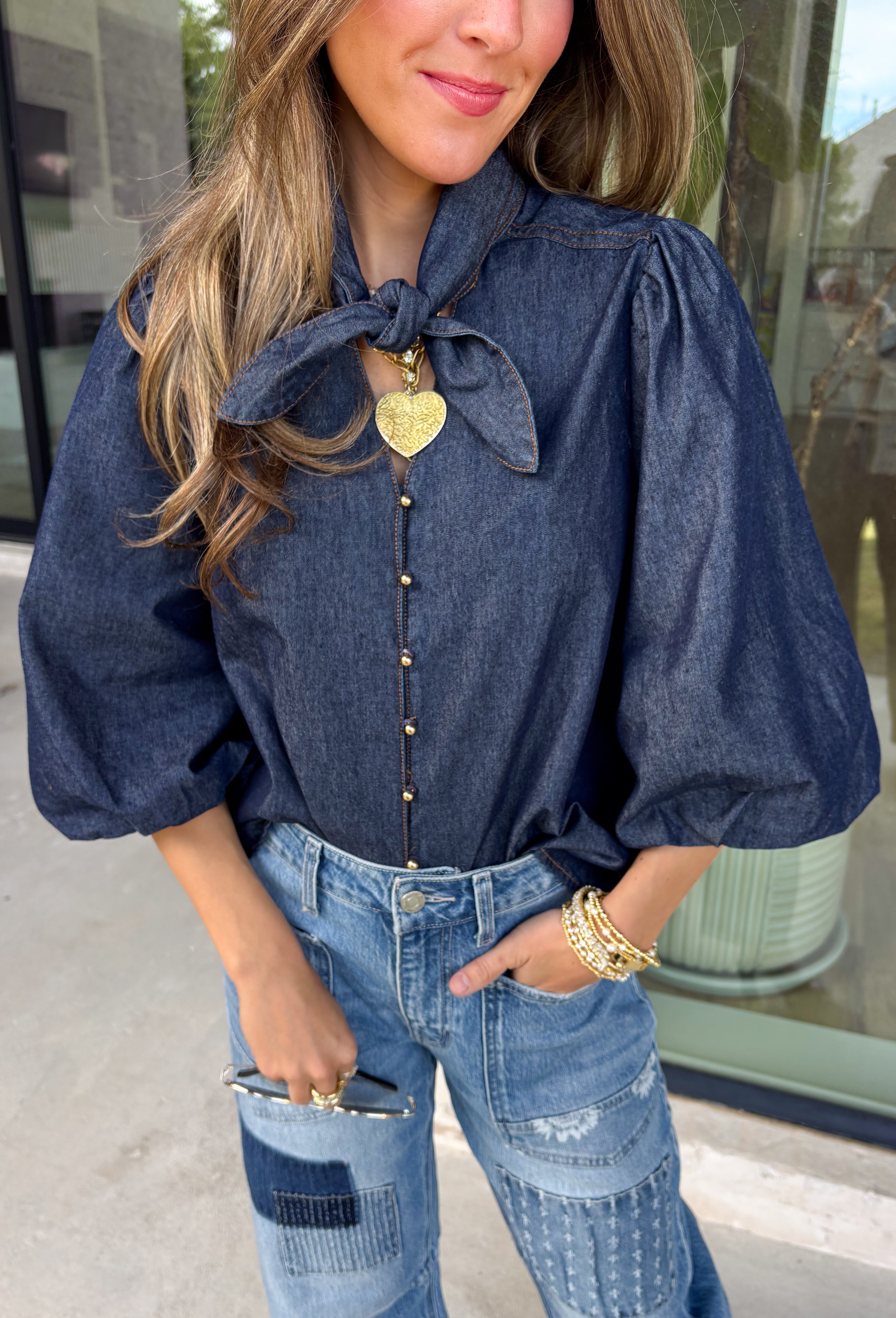 Give your denim collection a fresh twist with this dark wash blouse from Entro. The lightweight cotton fabric keeps it comfortable and easy to wear, while the rich dark wash feels right at home as fall transitions into winter. A high neckline with a self-tie detail adds a pretty focal point, complemented by gold button detailing along the front. The ¾ puff sleeves bring a little volume to the silhouette without making it feel overly dressy. 