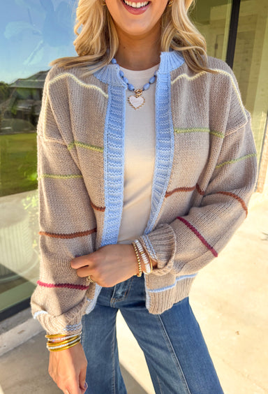 Cozy, colorful, and perfect for layering—this tan knit cardigan is a must-have for your fall wardrobe. Designed with bold stripes in burgundy, brown, green, light yellow, and light blue, it’s finished with light blue trim along the neckline and edges for a playful pop. The soft knit makes it warm without being heavy, giving you the ideal piece to throw over basics or style with denim and dresses alike.